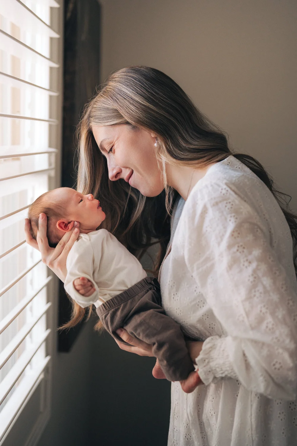Mom by the window holding her newborn close, both looking at each other, Hunter family in-home session Rancho Mission Viejo