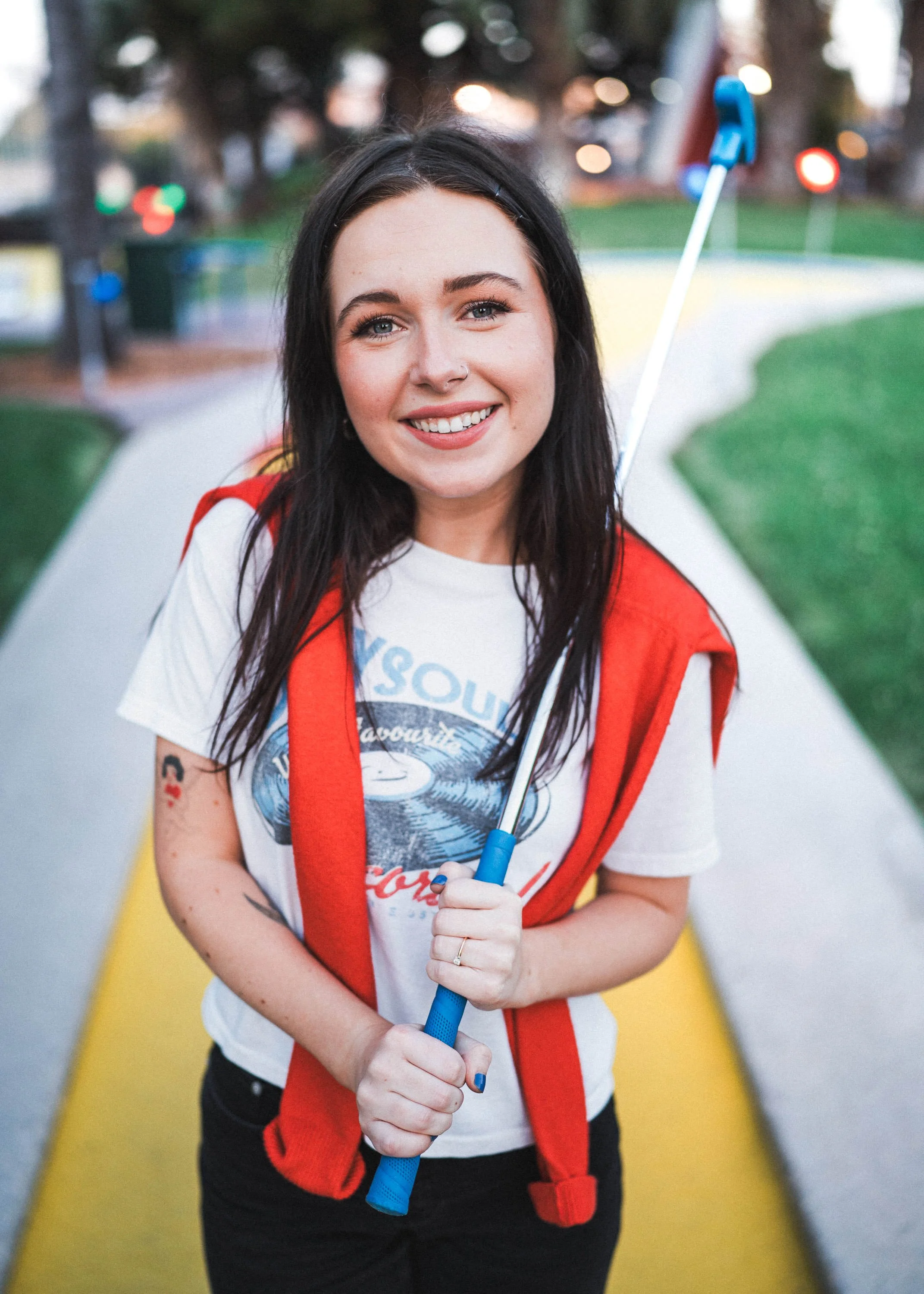 A young woman with dark hair, wearing a white graphic t-shirt and a red vest, smiles while holding a selfie stick outdoors on a yellow pathway in a park.