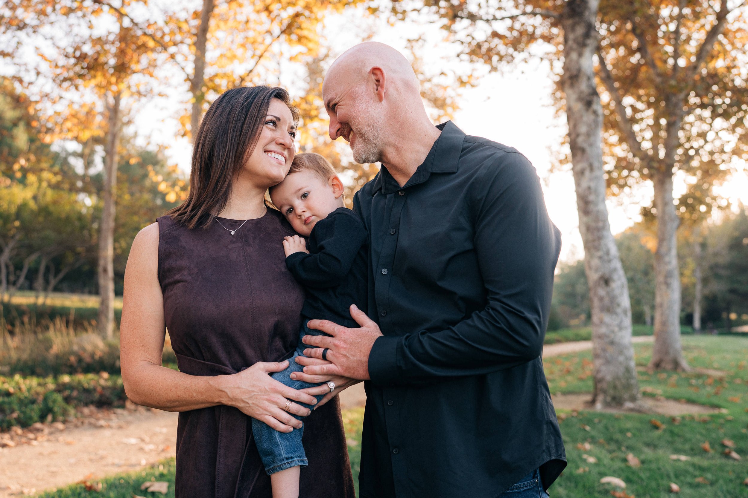 A family of three outdoors during autumn, with a woman holding a young child on her hip, and a man leaning in close, all smiling at each other amid trees with fall foliage.