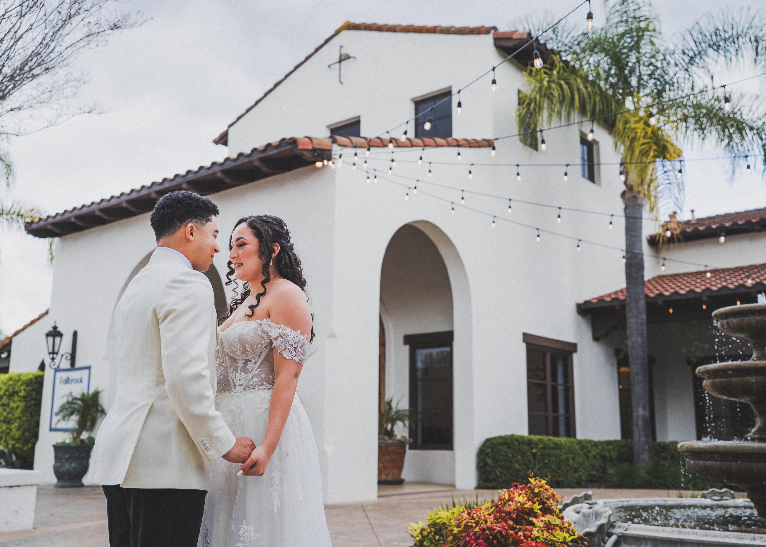 A bride and groom holding hands and gazing at each other outside a white building with string lights hanging above. The bride has dark curly hair and is wearing a white gown with lace details. The groom is dressed in a white suit jacket. There are po