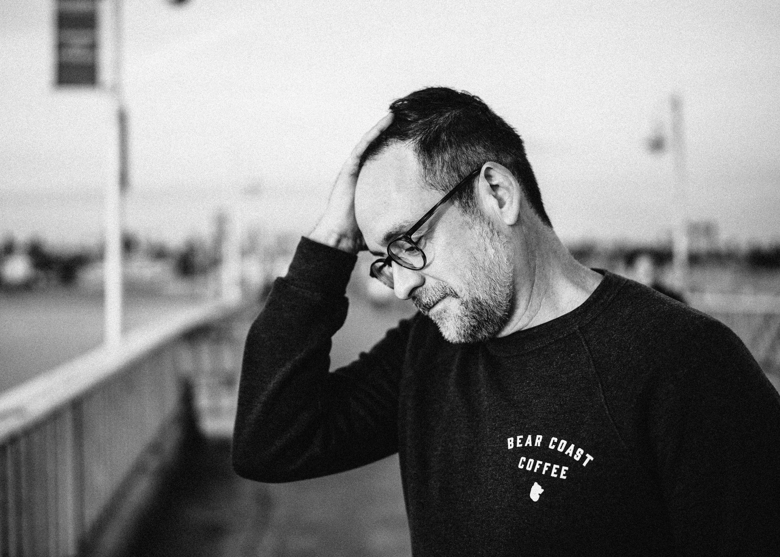 A black and white photo of a middle-aged man with glasses and a short beard, holding his head with one hand and looking downward, standing outdoors near a railing, with an urban setting in the blurry background.