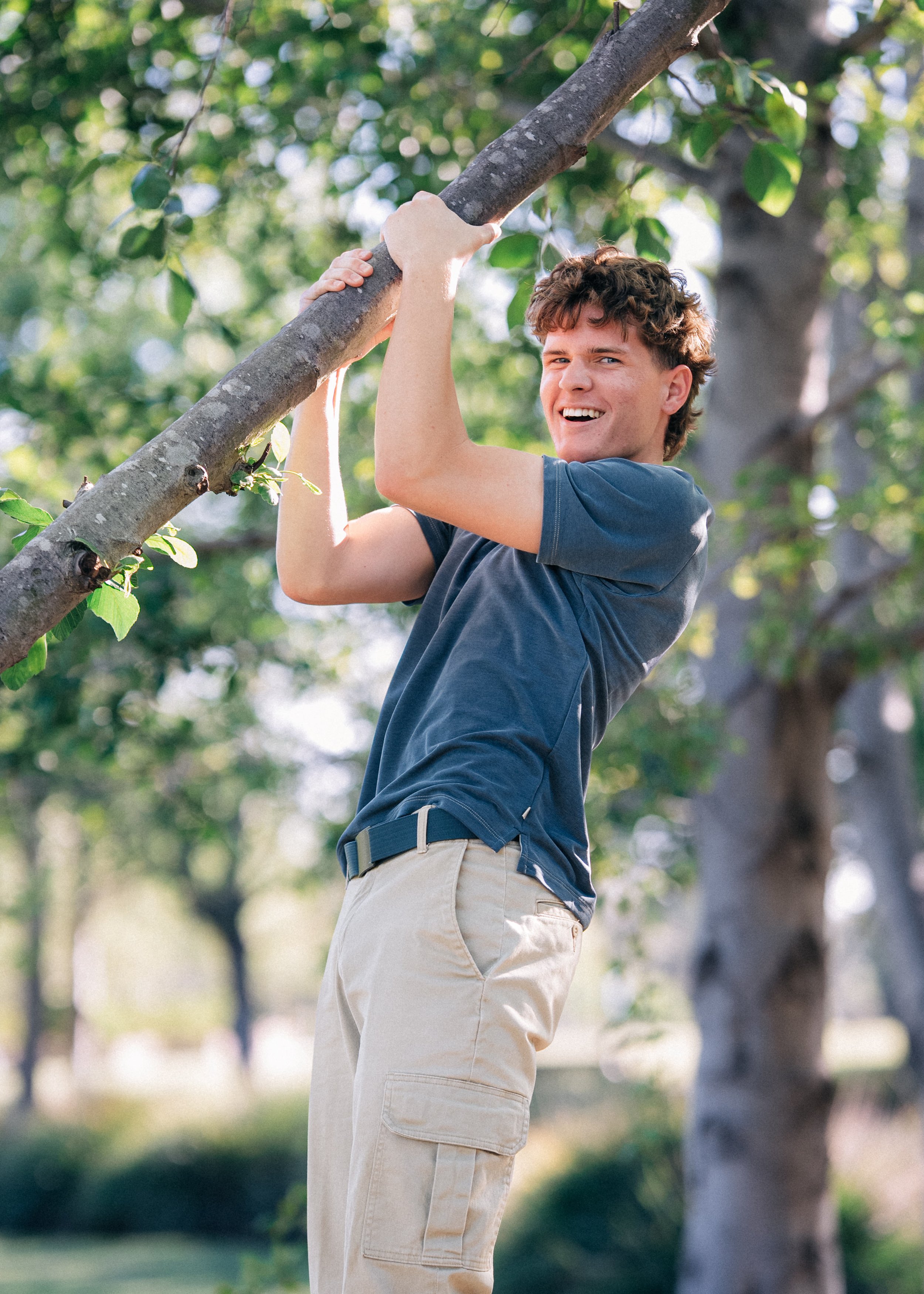 A young man with curly brown hair in a blue t-shirt and khaki shorts is climbing and hanging from a tree branch outdoors in a park, smiling.