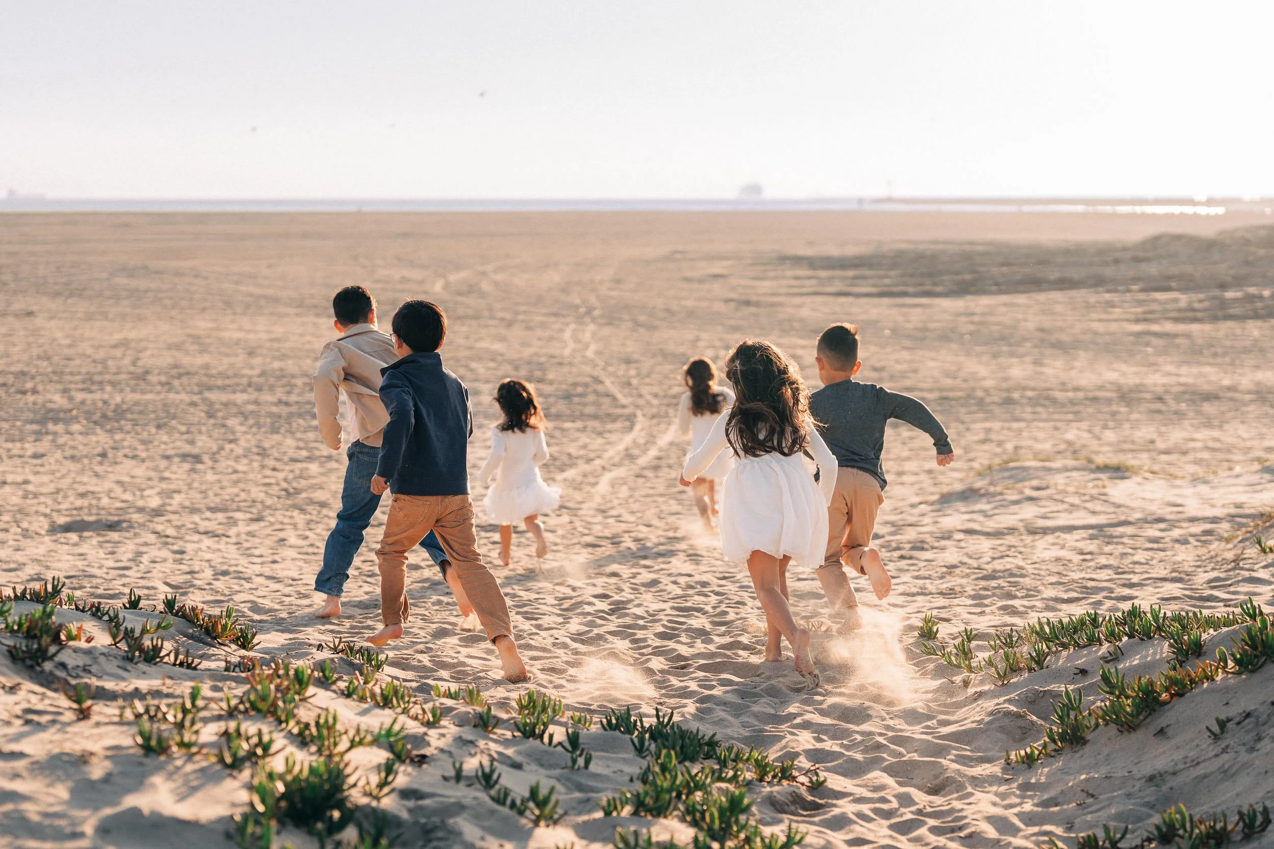 Children running on sandy beach during daytime with low sun, behind vegetated dunes.