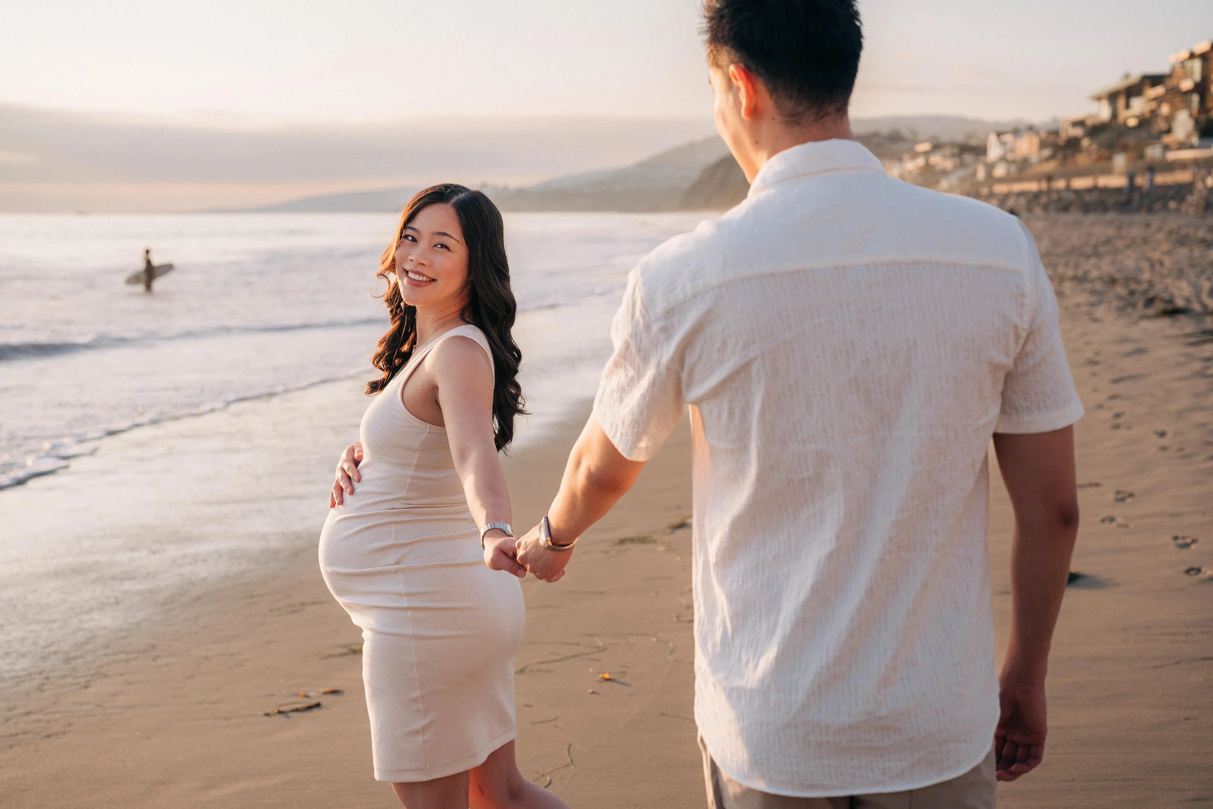 A smiling pregnant woman holding hands with a man on the beach during sunset, with the ocean and hills in the background.
