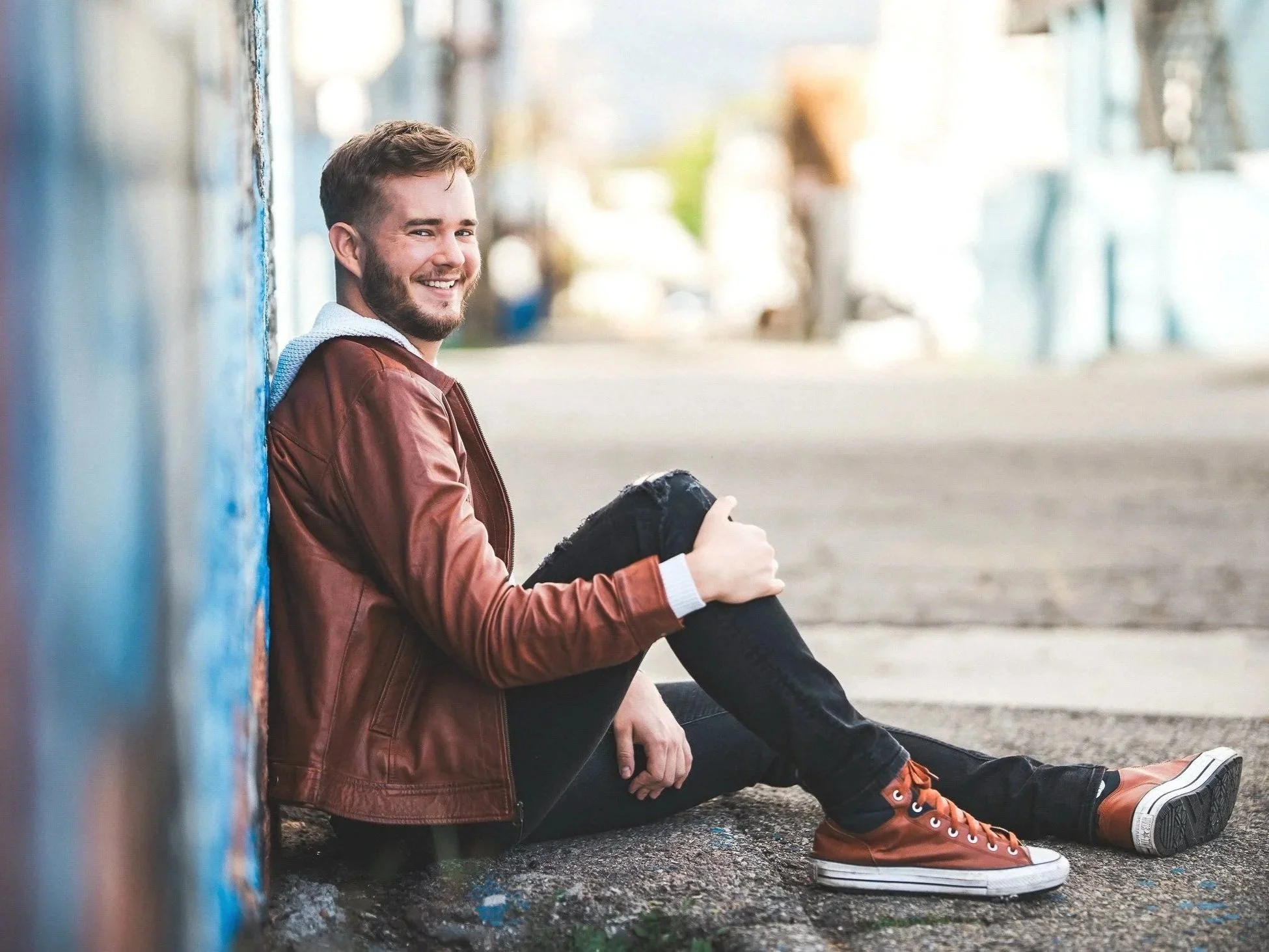 A young man with a beard is sitting on the ground against a blue wall, smiling, in an urban setting. He is wearing a brown leather jacket, black pants, and orange high-top sneakers.