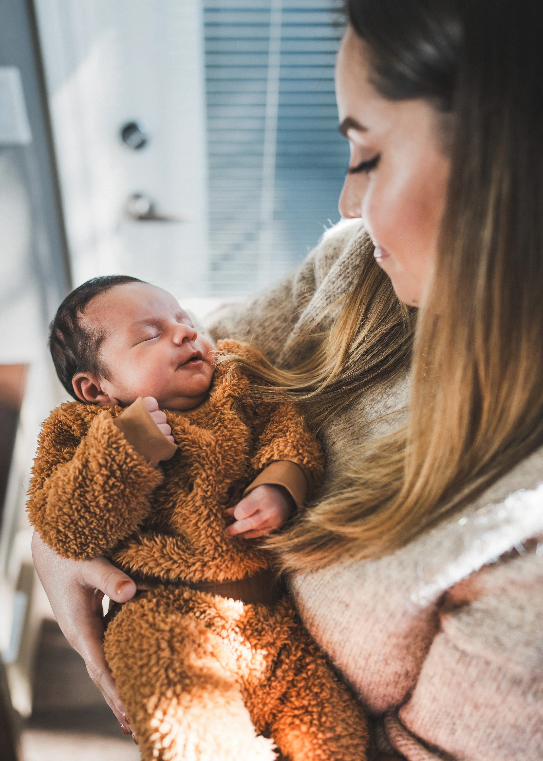 A woman with long hair is holding a sleeping baby in a brown fleece outfit close to her chest, inside a room with blinds on the window.
