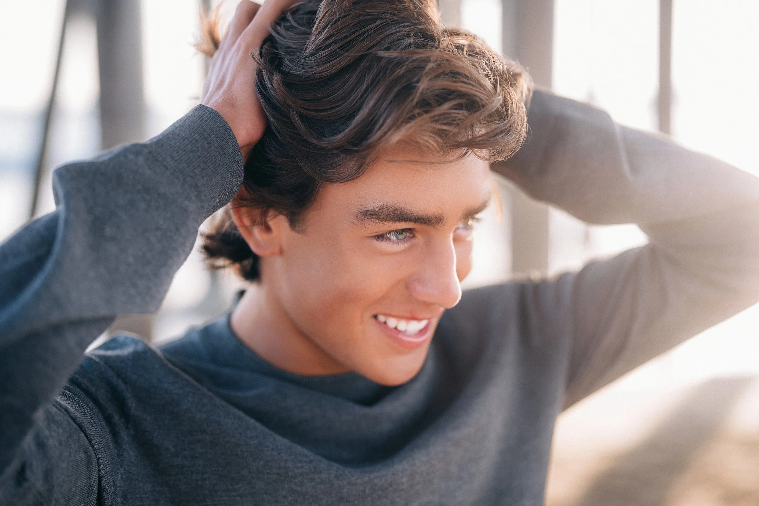A young man with brown hair and blue eyes smiling, touching his head with both hands, wearing a dark gray long-sleeve shirt.