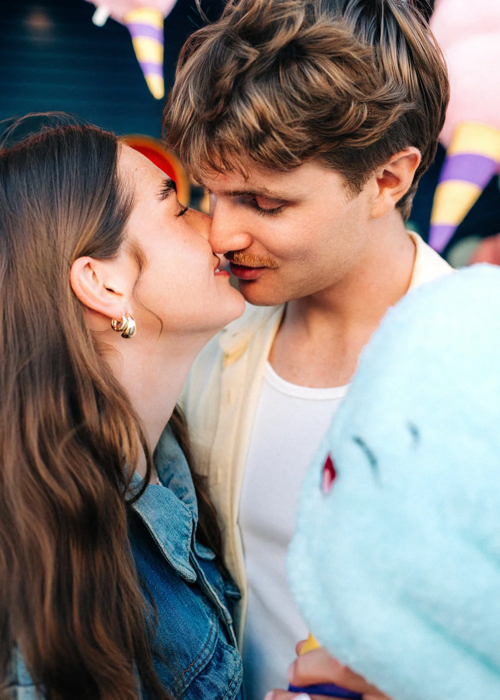 Quinn and Parker sharing a kiss in front of colorful carnival plush prizes at Belmont Park, San Diego
