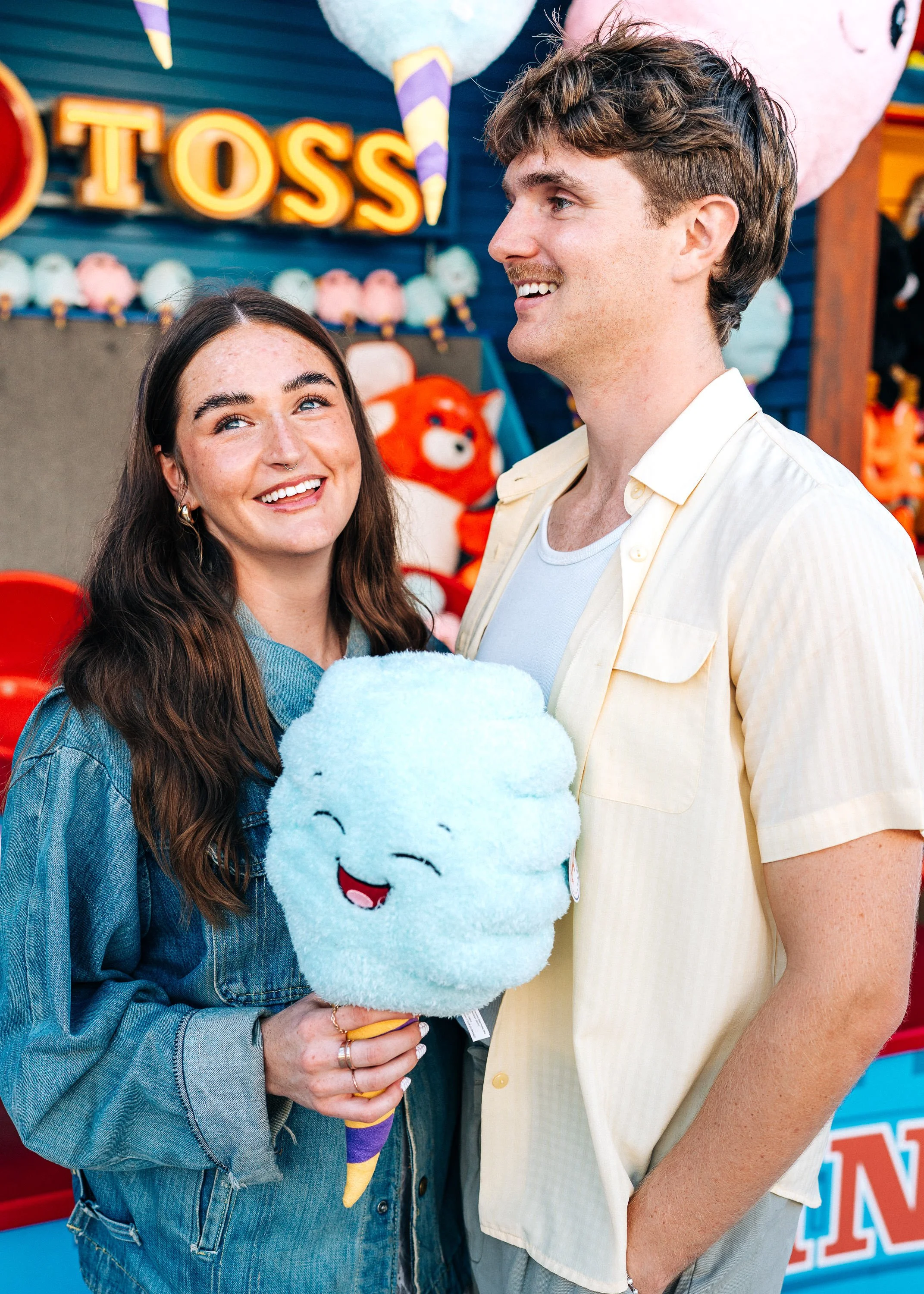 A young woman and man smiling at each other at an amusement arcade, with arcade games and stuffed animal prizes in the background. The woman is holding a plush cotton candy with a happy face.