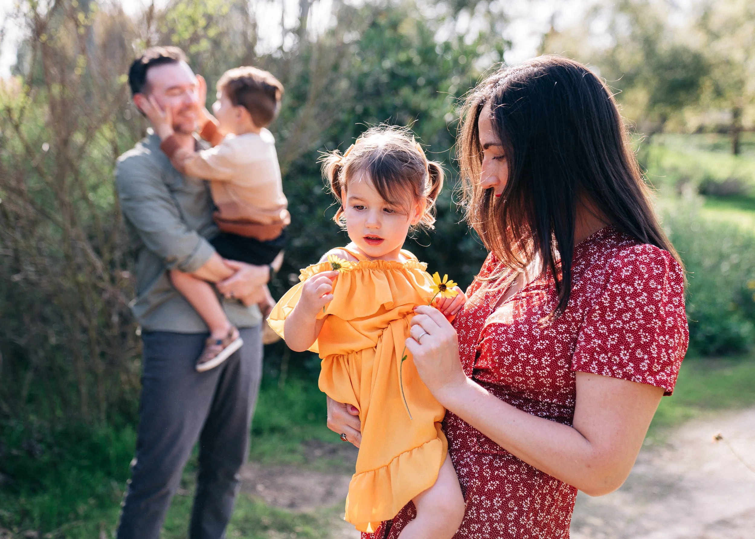 A woman in a red patterned dress holding a young girl in an orange dress, both holding yellow flowers, outdoors with a blurred background of trees and a man holding a boy in the background.