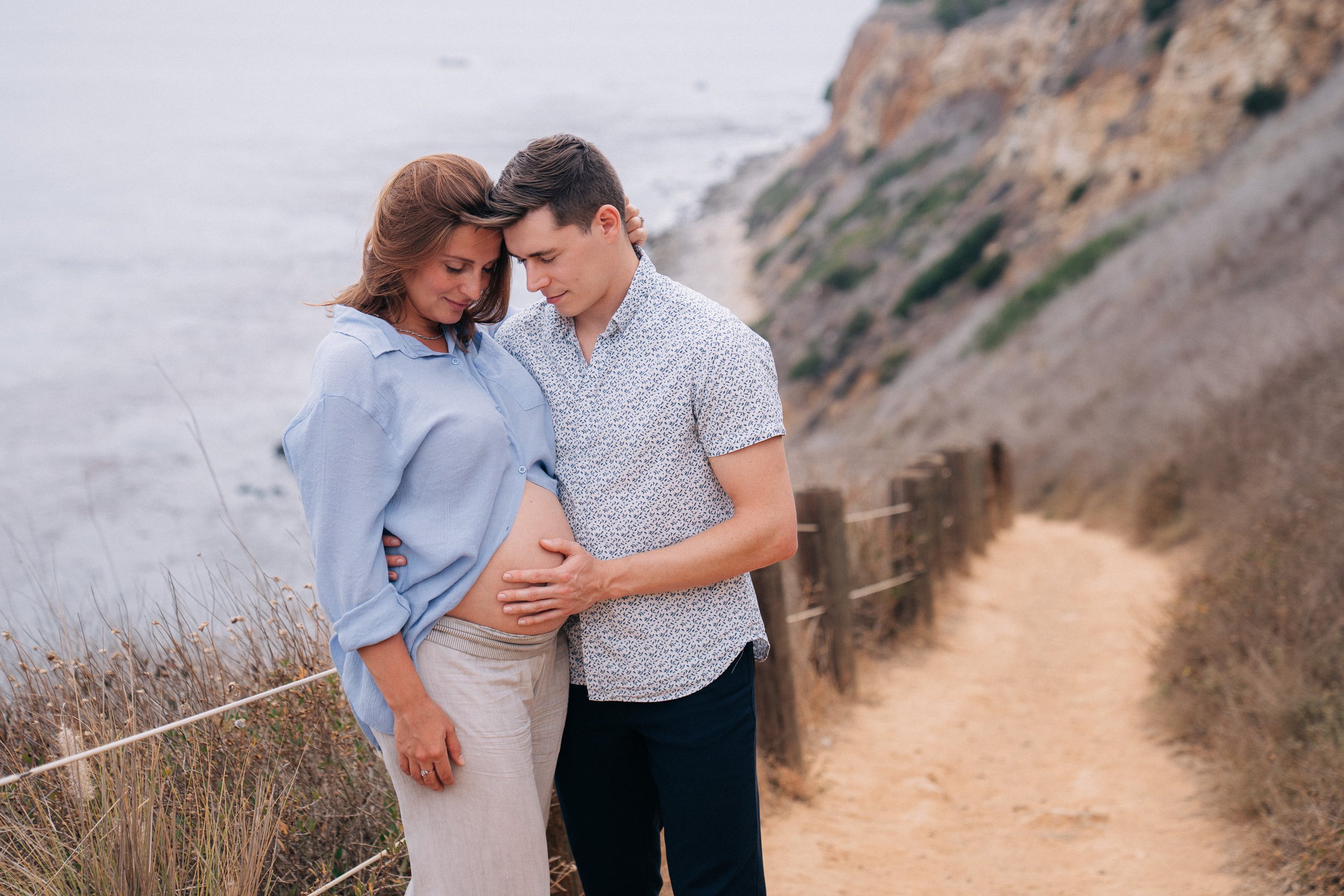 A couple on a nature trail with a cliffside view, the pregnant woman and man are touching her belly in an affectionate pose.