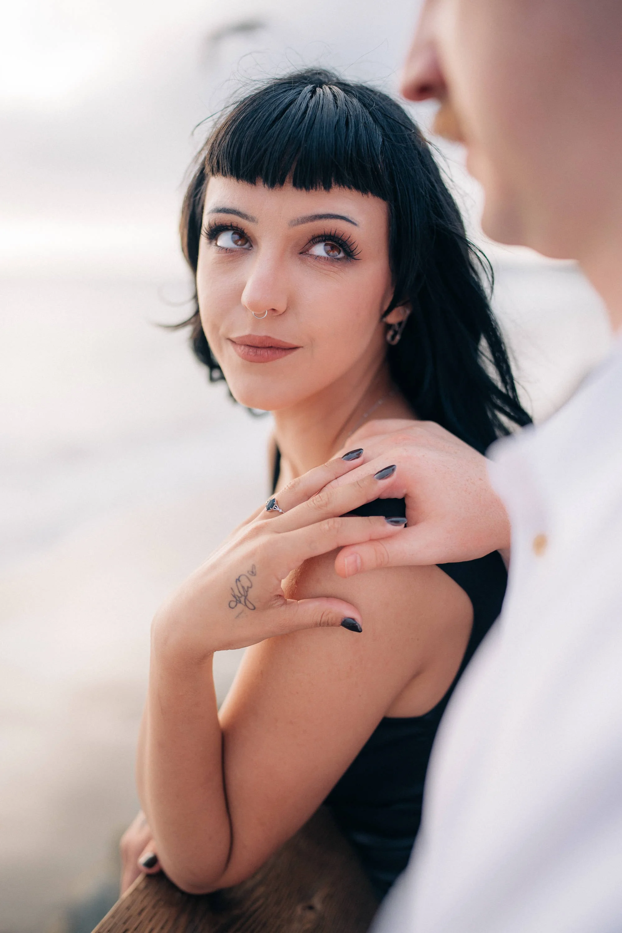 A woman with short black hair, wearing makeup, and a septum piercing, looks at a person partially visible on the right. She is holding her shoulder with her left hand, which has a tattoo and dark nail polish, and is outdoors on a cloudy day.