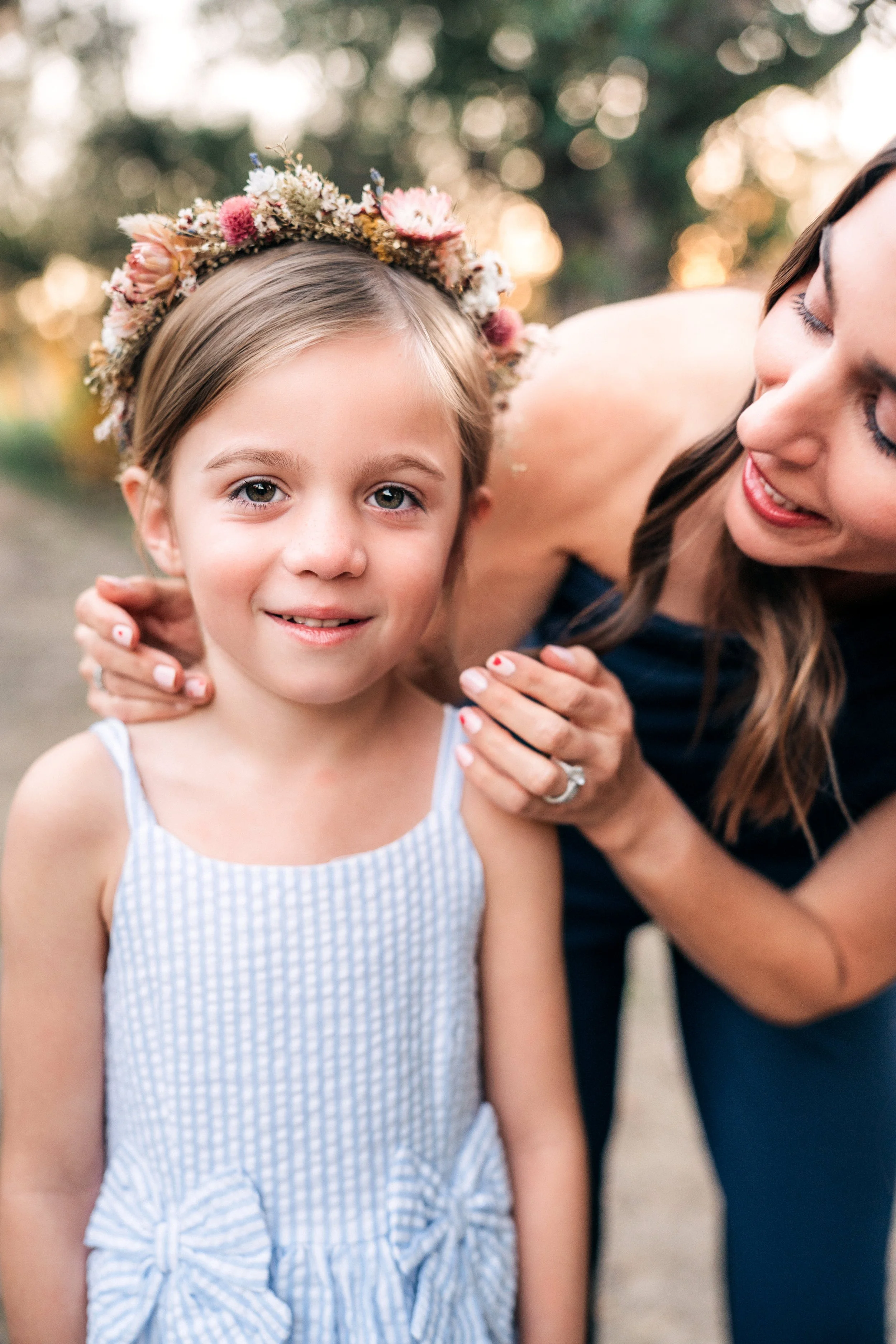 A young girl with blue eyes and a floral crown smiles as an adult woman, possibly her mother, gently holds her shoulders and smiles.
