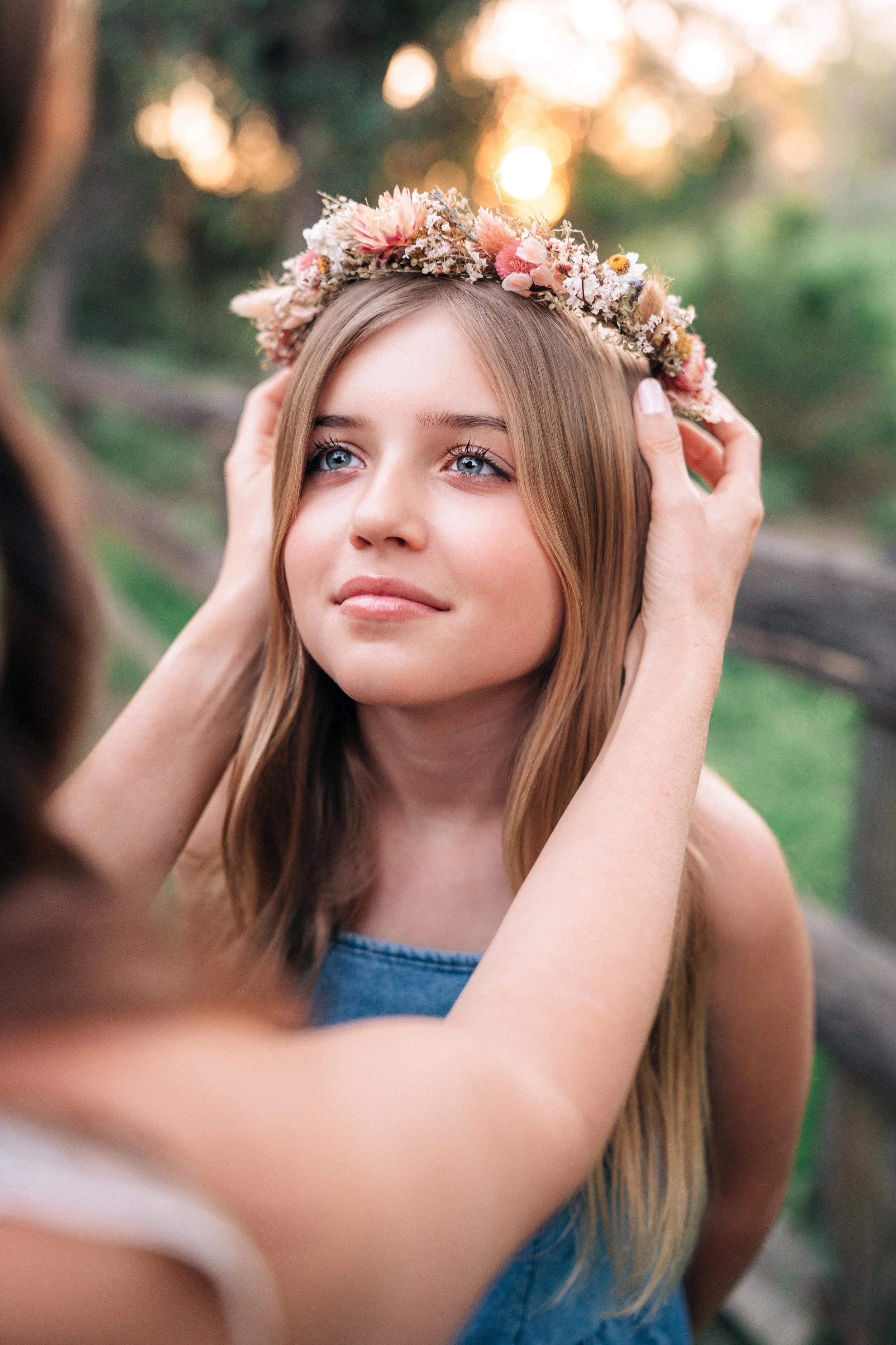 A young woman with long, light brown hair and blue eyes is being helped to put a floral crown on her head by another person. The background shows a natural outdoor setting with trees and sunlight.