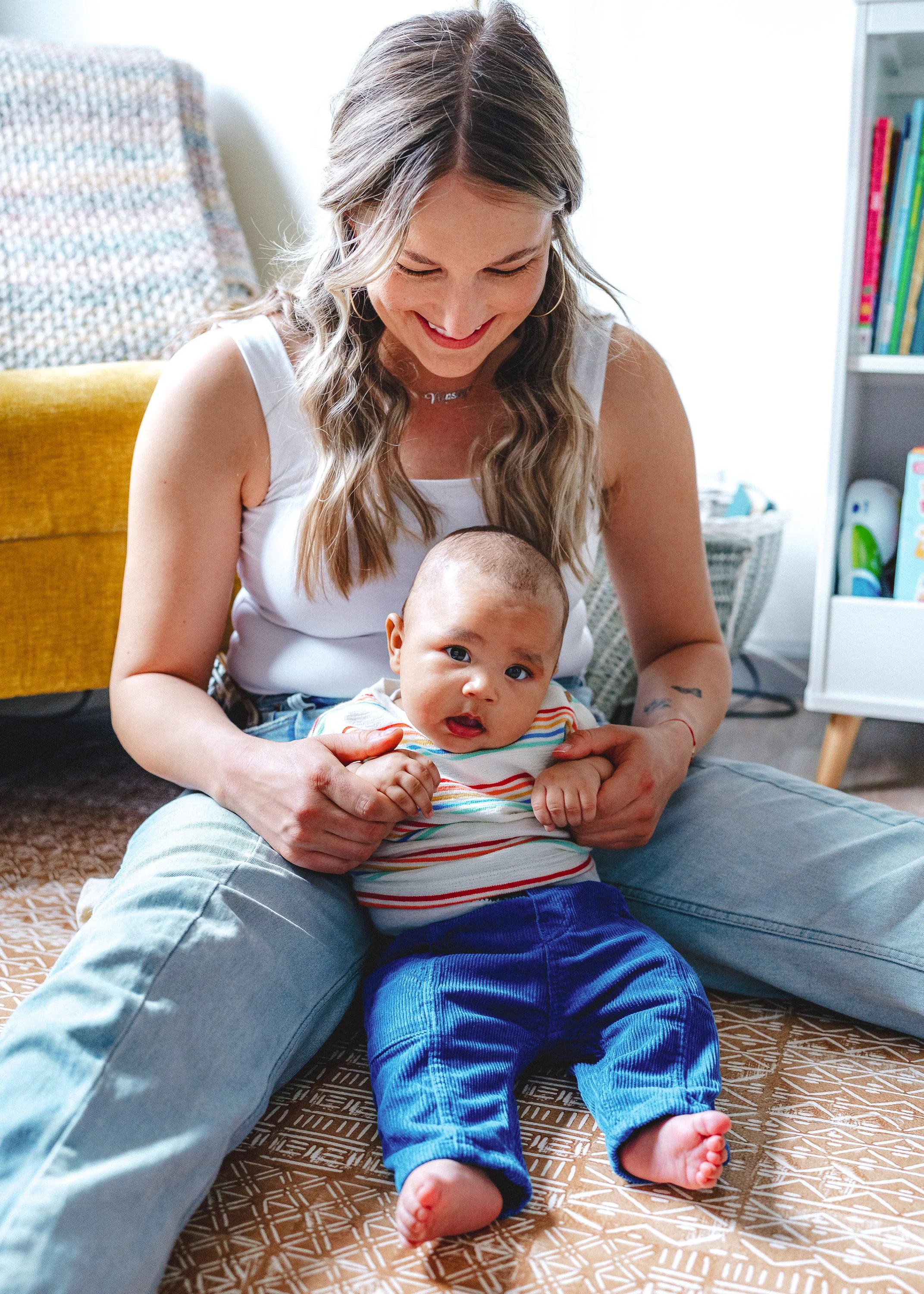 A woman with wavy, light brown hair and a white sleeveless top sitting on the floor, smiling and holding a young baby with short dark hair and wearing a striped shirt and blue pants. They are indoors with a yellow couch and a bookshelf in the backgro