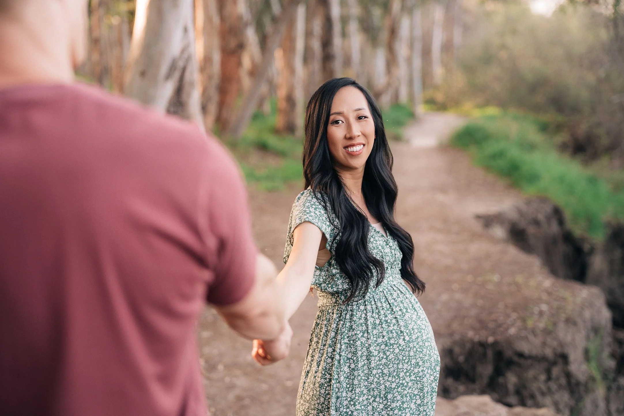 A pregnant woman with long black hair, wearing a green floral dress, standing on a dirt path in a forest, is holding hands with someone off-camera and looking back with a smile.
