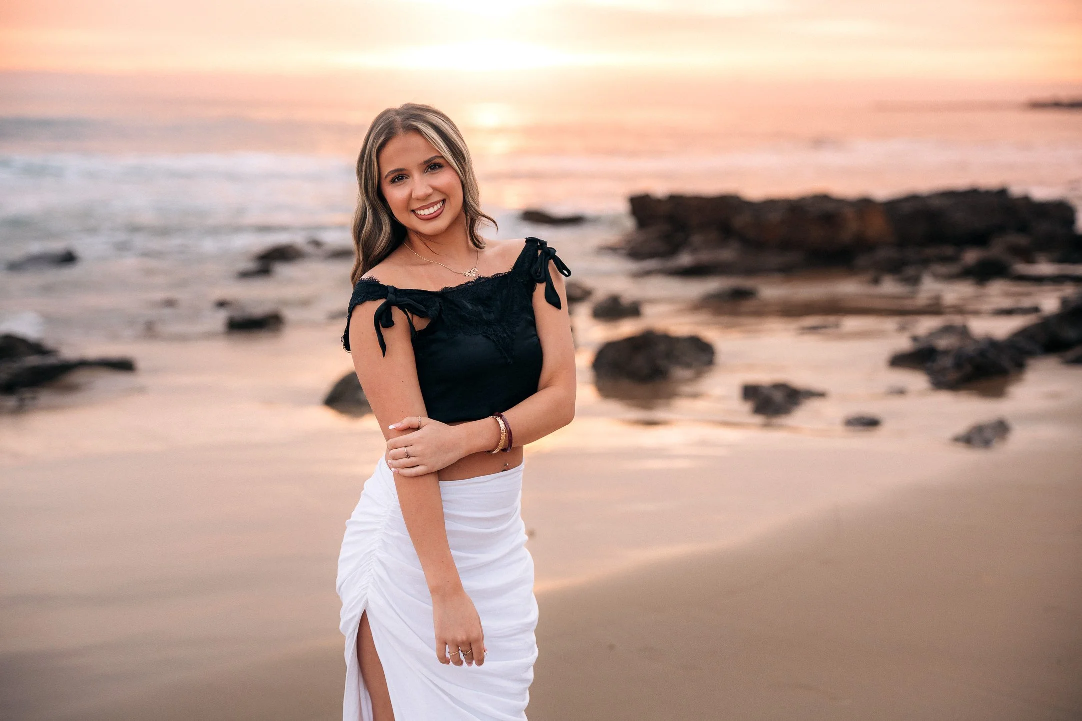 A young woman smiling at the camera standing on a beach at sunset, wearing a black top with tie straps and a white skirt.