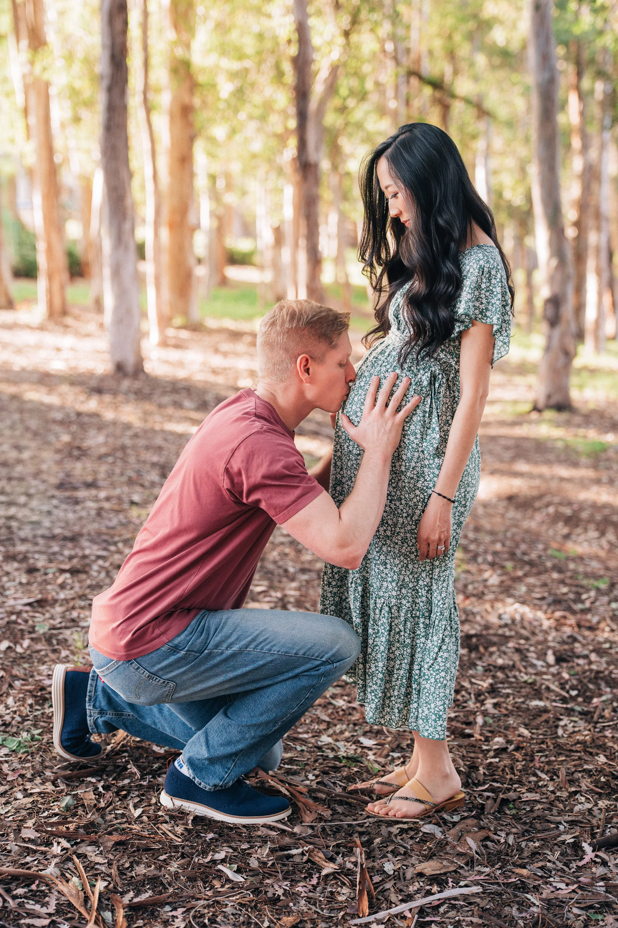 A man kneeling and kissing a pregnant woman's belly in a forest with sunlight filtering through trees.