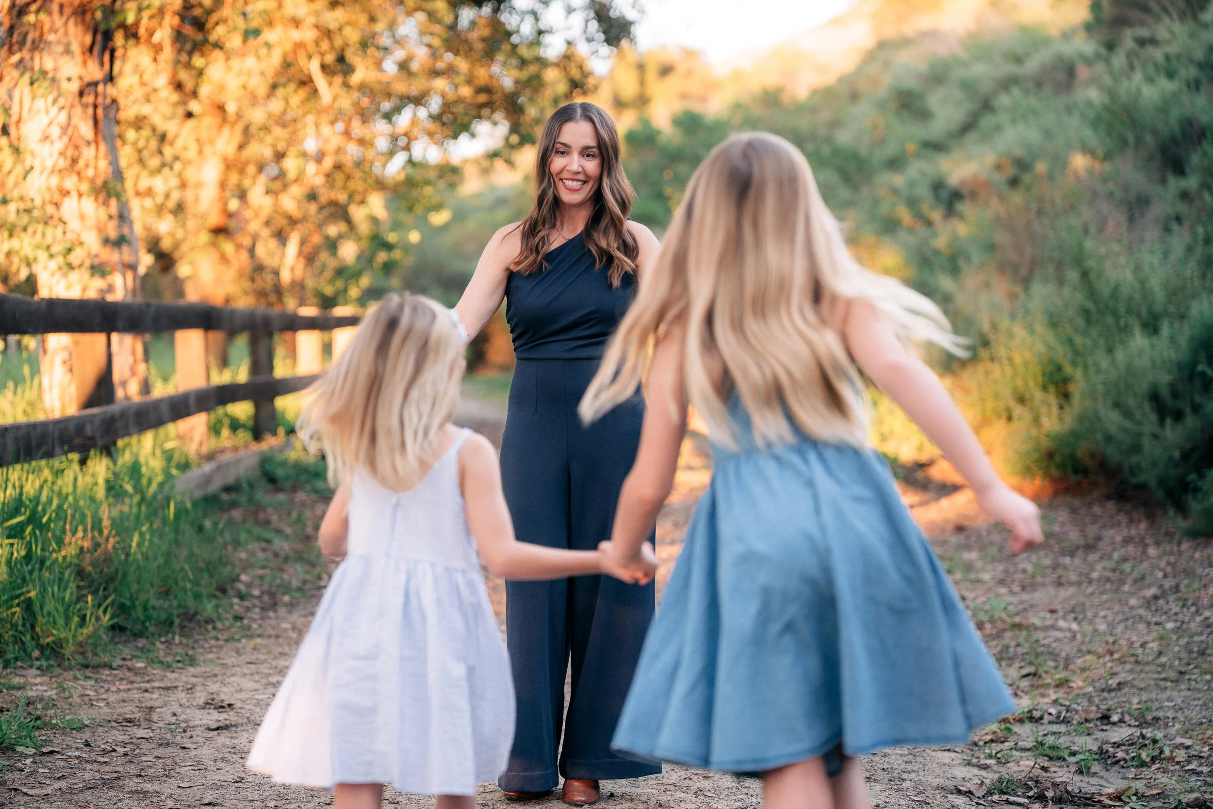 A woman in a dark jumpsuit playing with two young girls in summer dresses on a dirt path in a park during sunset.