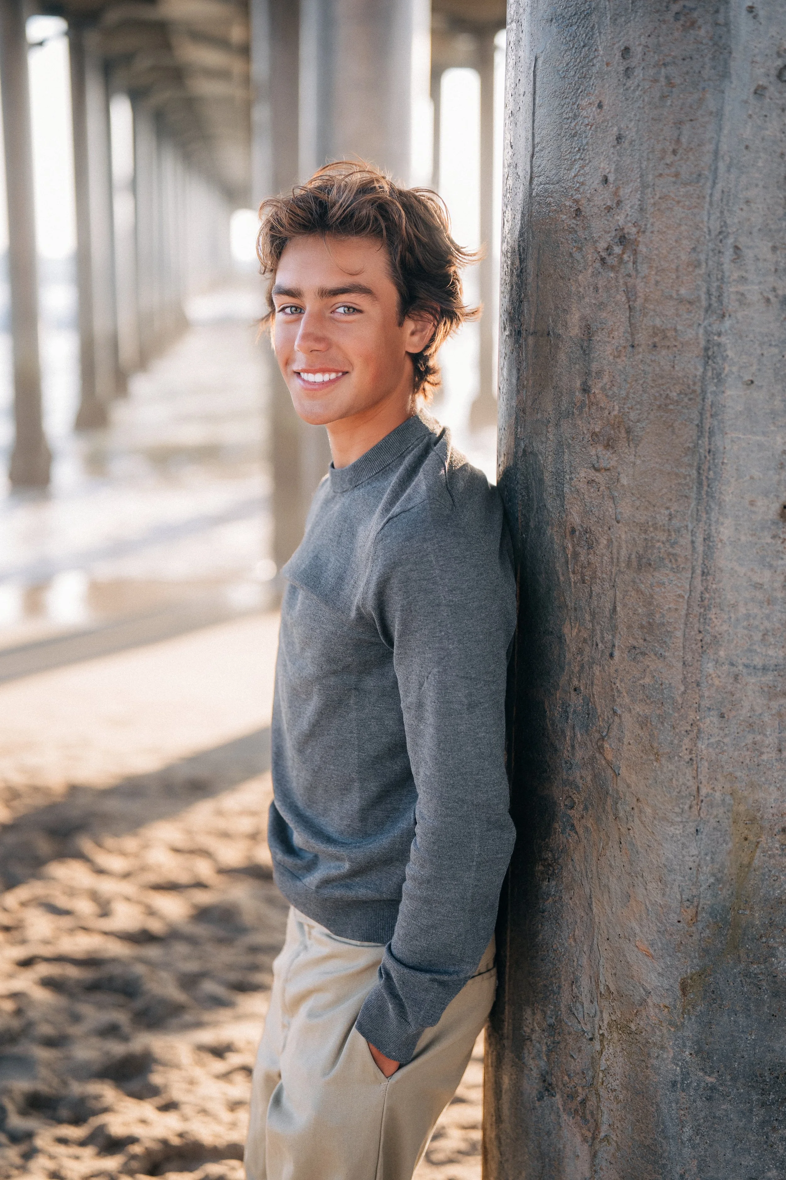A young man with wavy brown hair and a bright smile, leaning against a concrete pillar under a pier at the beach.