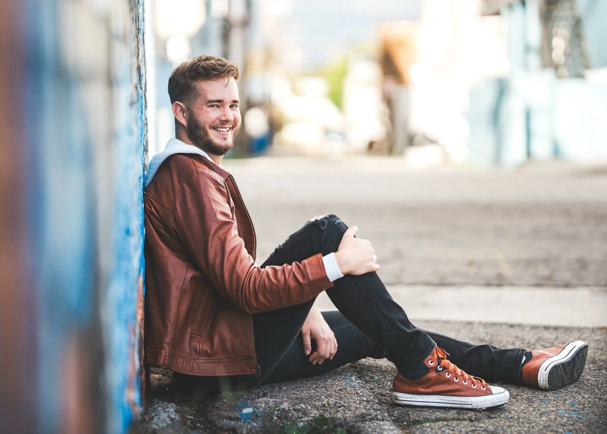 A young man with a beard sitting on the sidewalk, leaning against a graffiti-painted wall, smiling and looking at the camera.