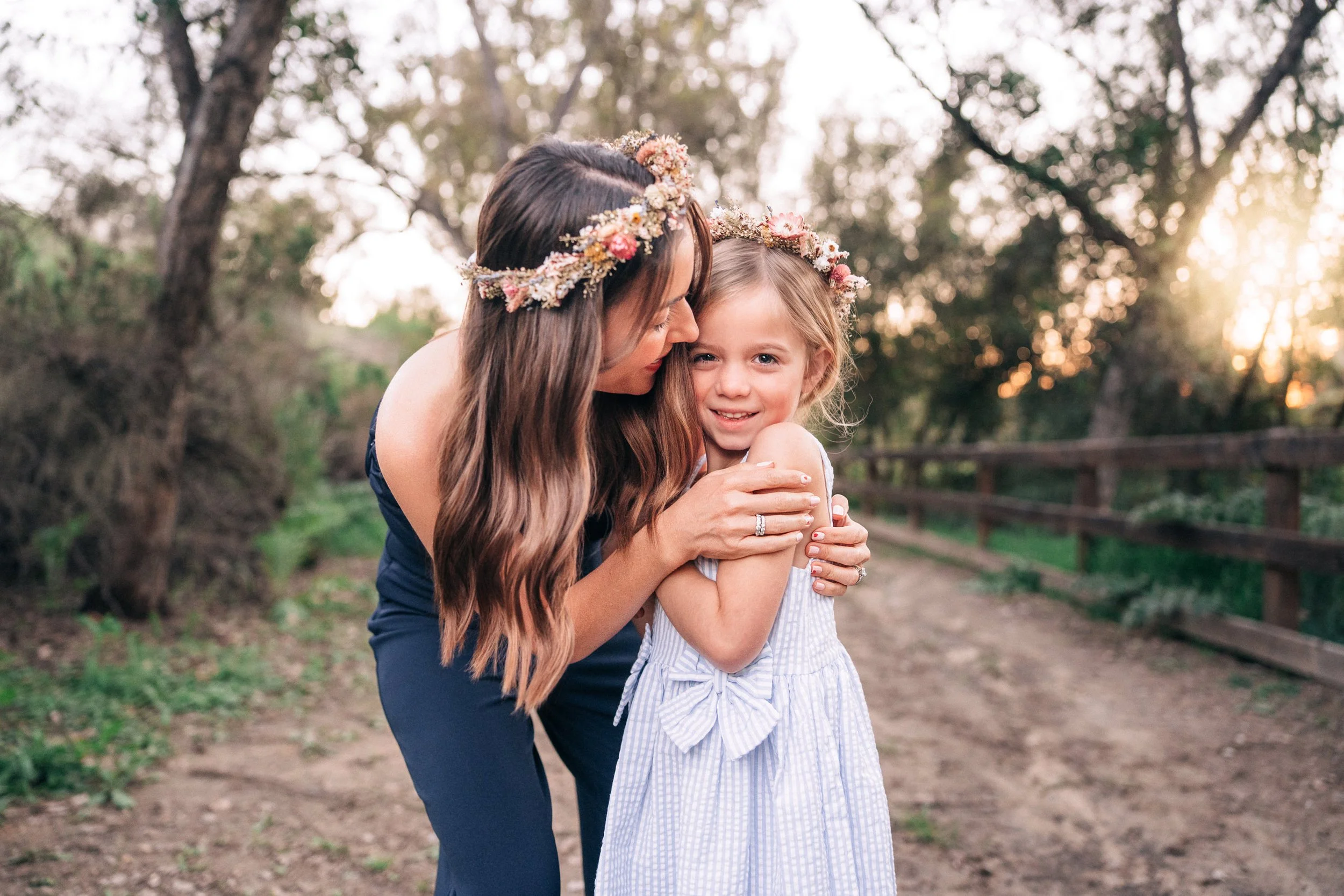 A woman and a young girl wearing floral crowns hugging each other outdoors during sunset.