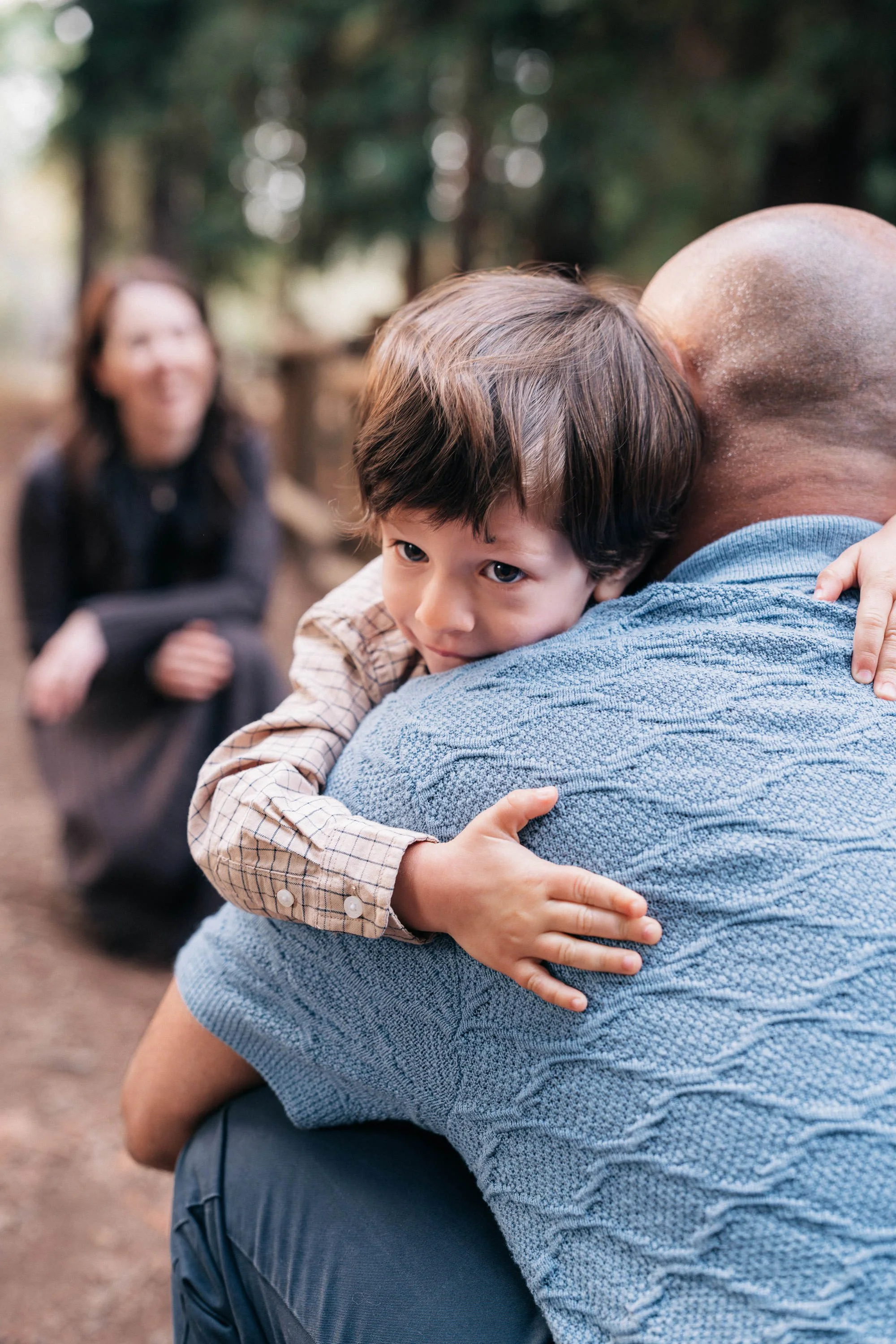 A young boy hugging an adult man outdoors, with a woman sitting in the blurred background.