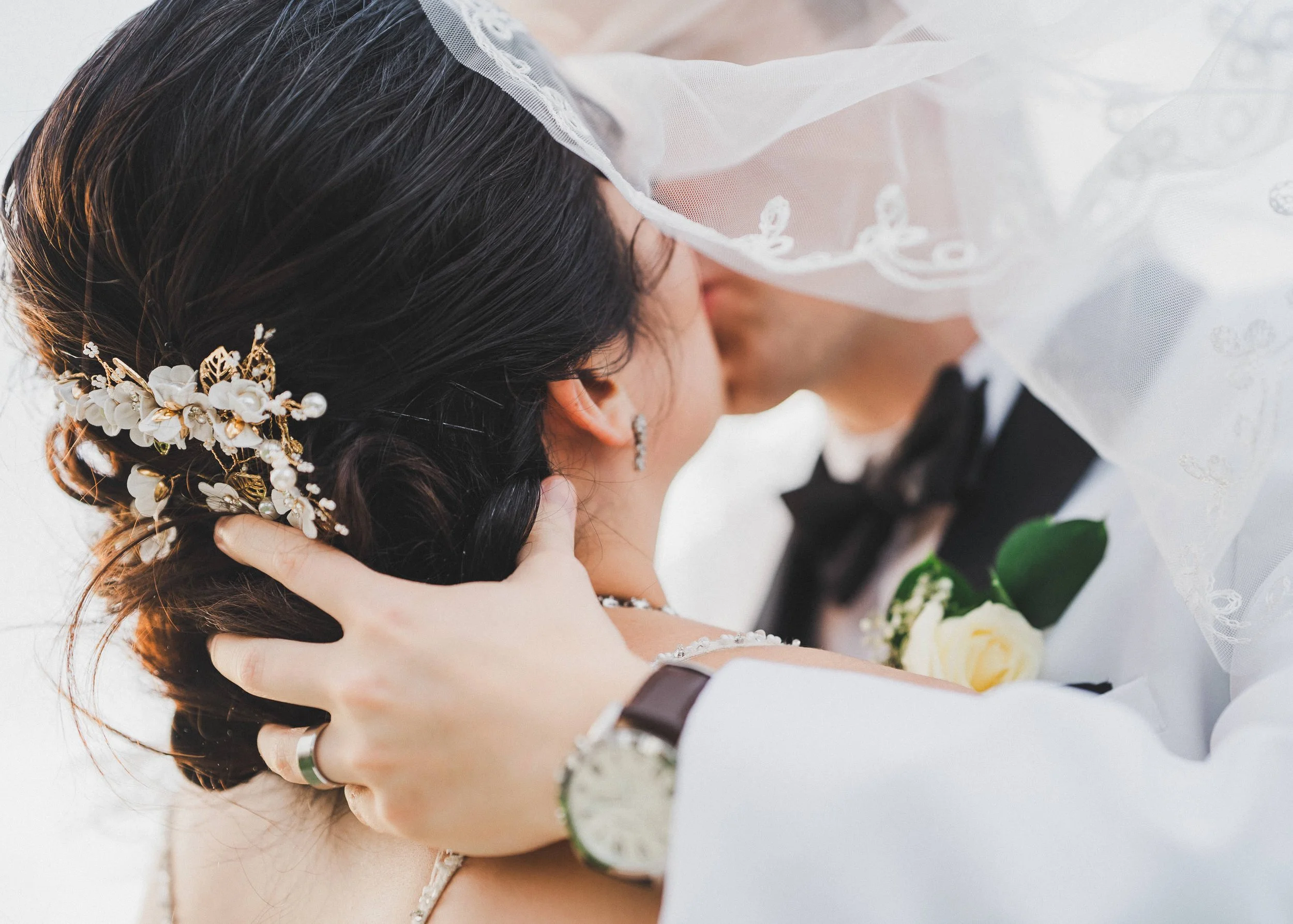 A bride and groom share a kiss, with the bride wearing a bridal headpiece and veil, and the groom in a tuxedo with a boutonniere.