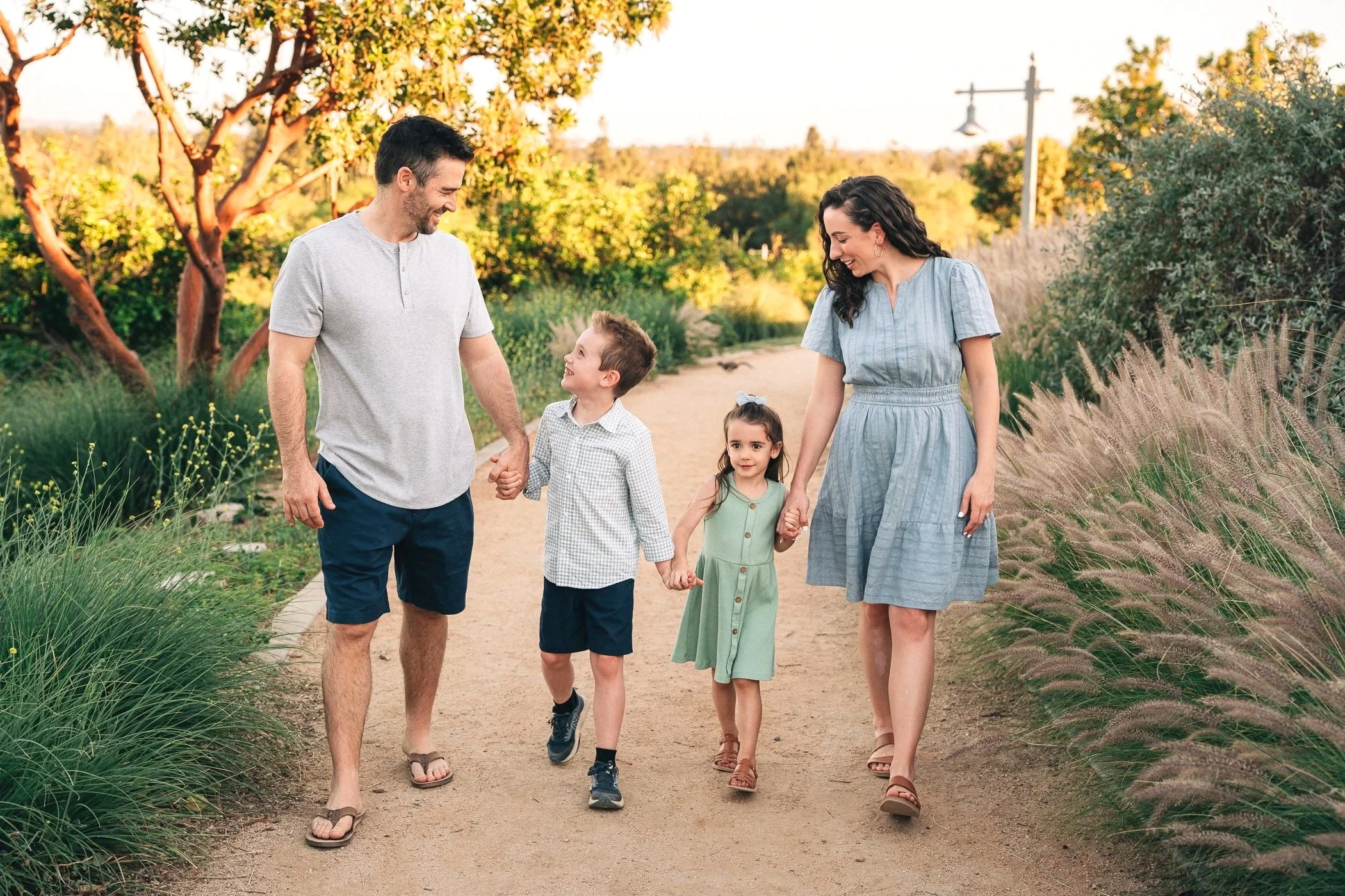 A family of five walking along a dirt path in a park during golden hour. The father is holding hands with a boy, and the mother is holding hands with a girl. Everyone is smiling and looking at each other. The background features green trees, bushes, and a streetlamp.