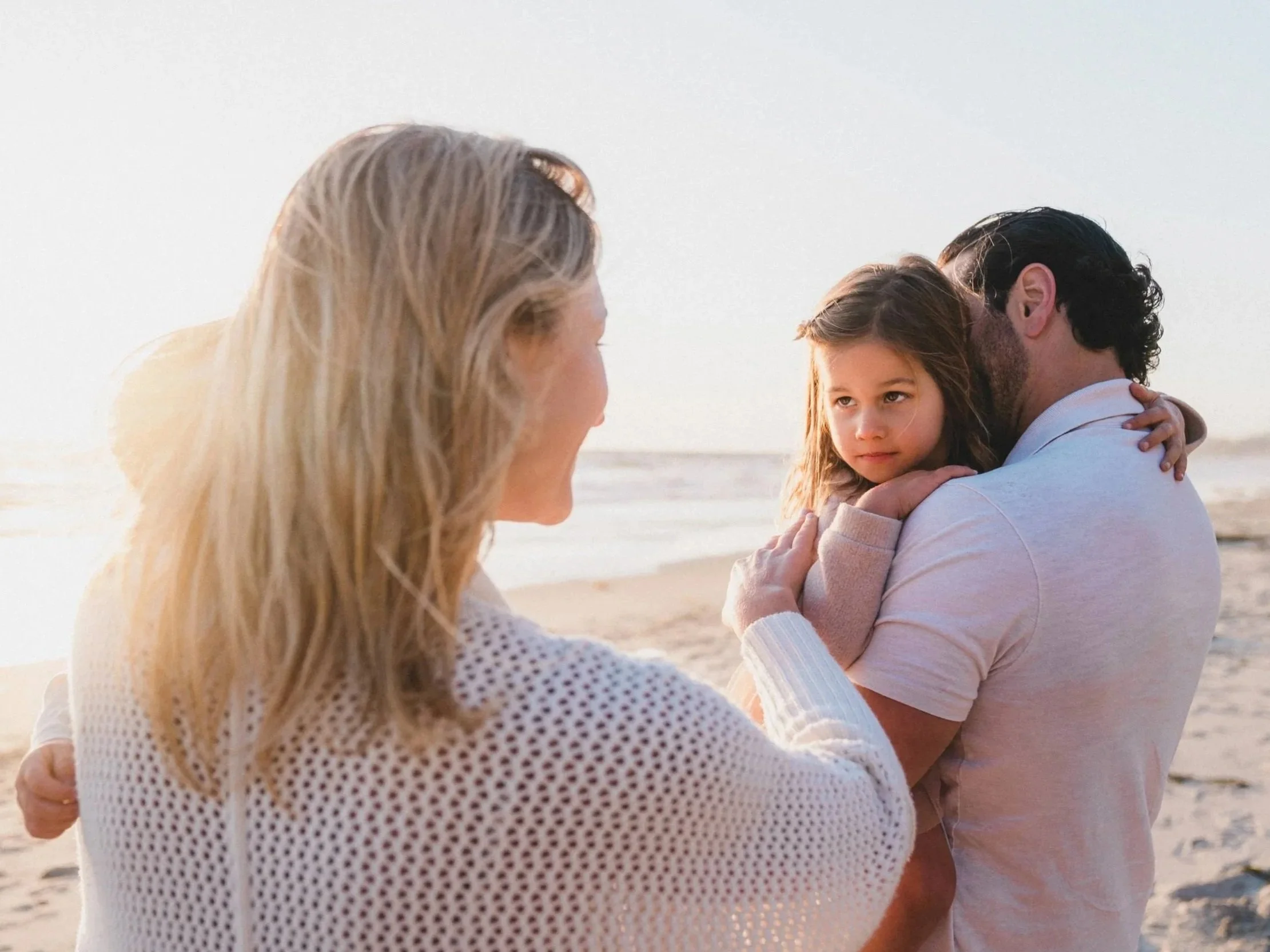 A family of three on the beach during sunset, with a woman facing away, a man holding a young girl, all embracing each other.