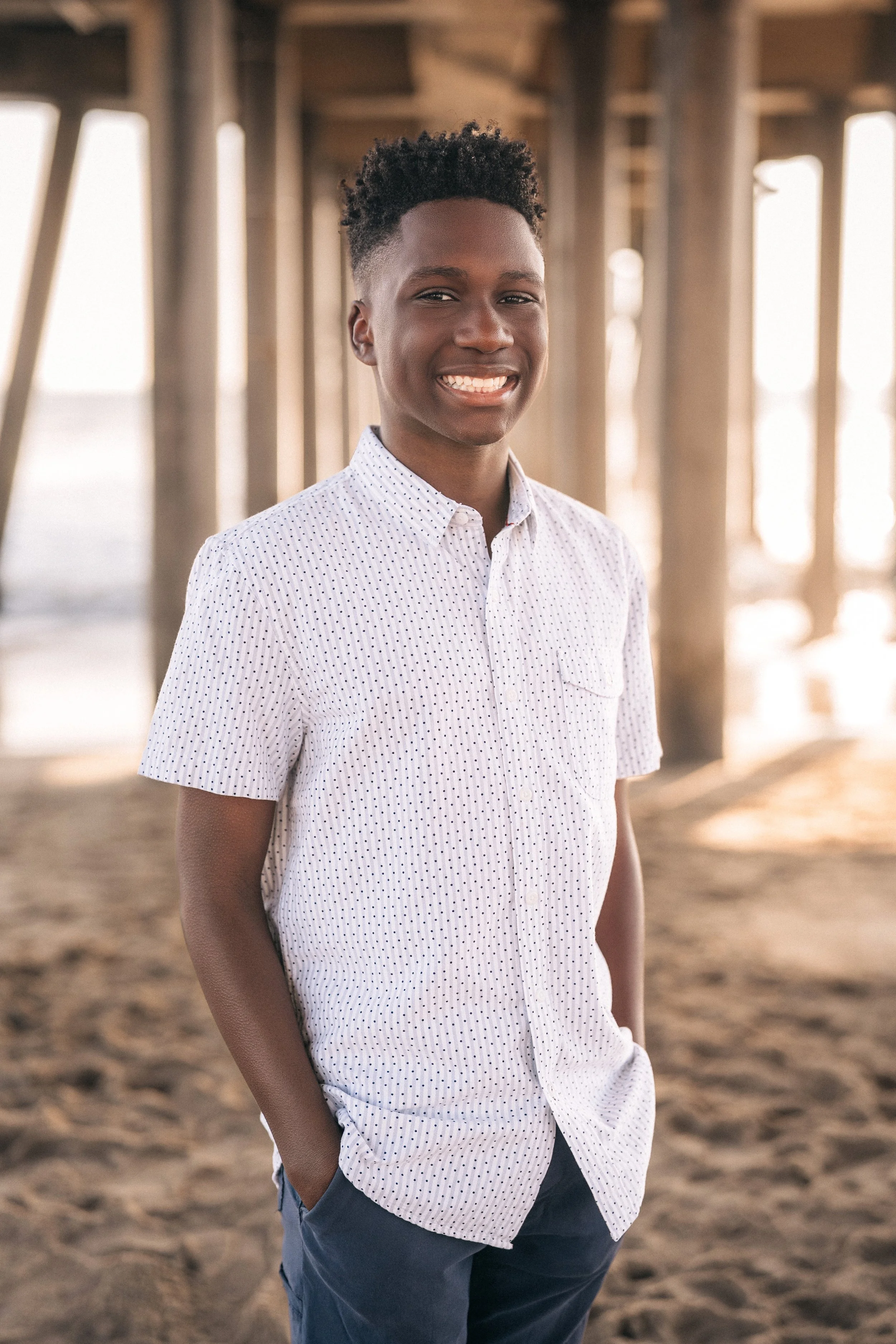 A young man smiling, dressed in a white, dotted short-sleeve button-up shirt and blue pants, standing on a sandy beach under a pier with wooden beams and sunlight filtering through.
