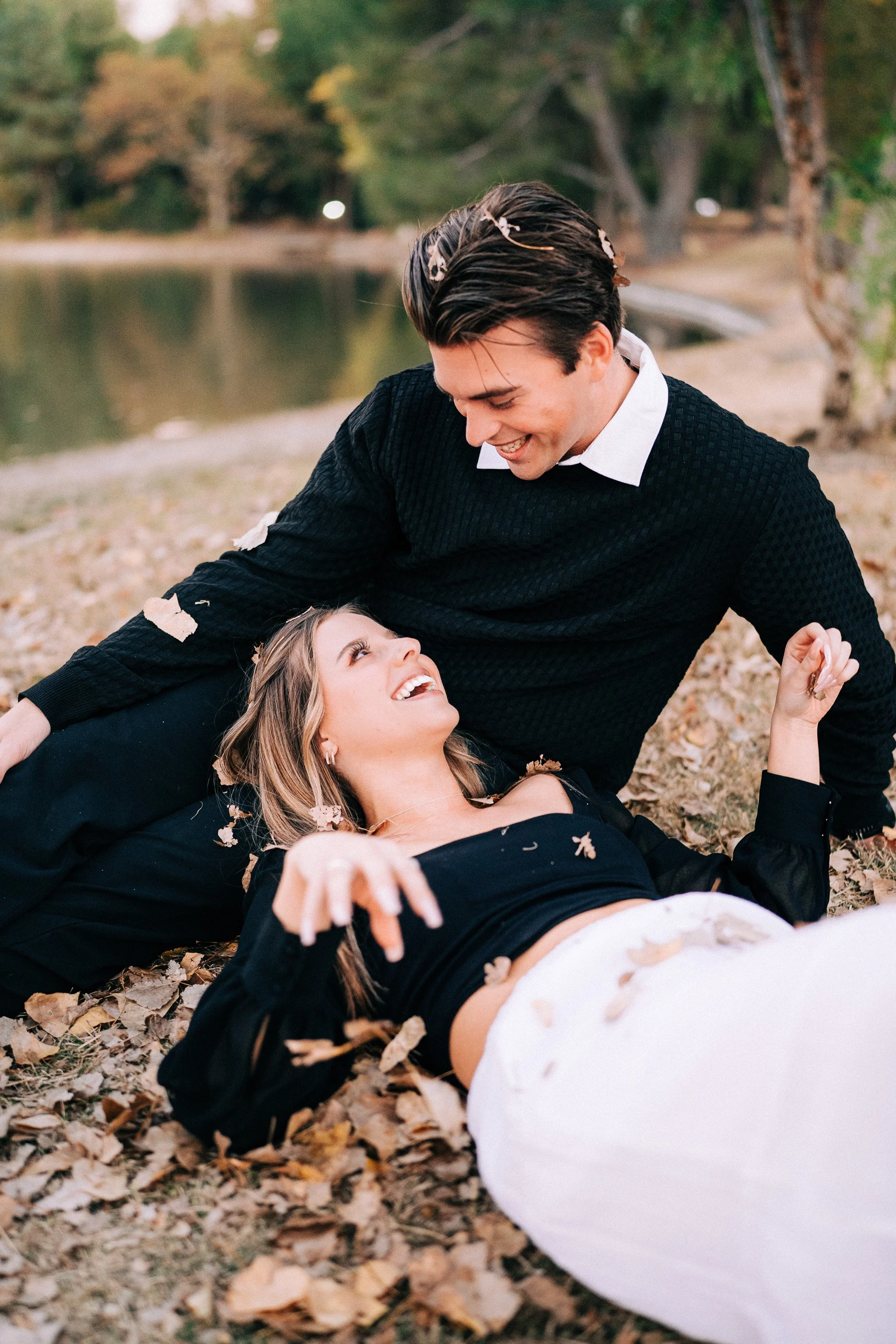 A young couple lying on the ground covered with autumn leaves near a lake, smiling at each other in a park.