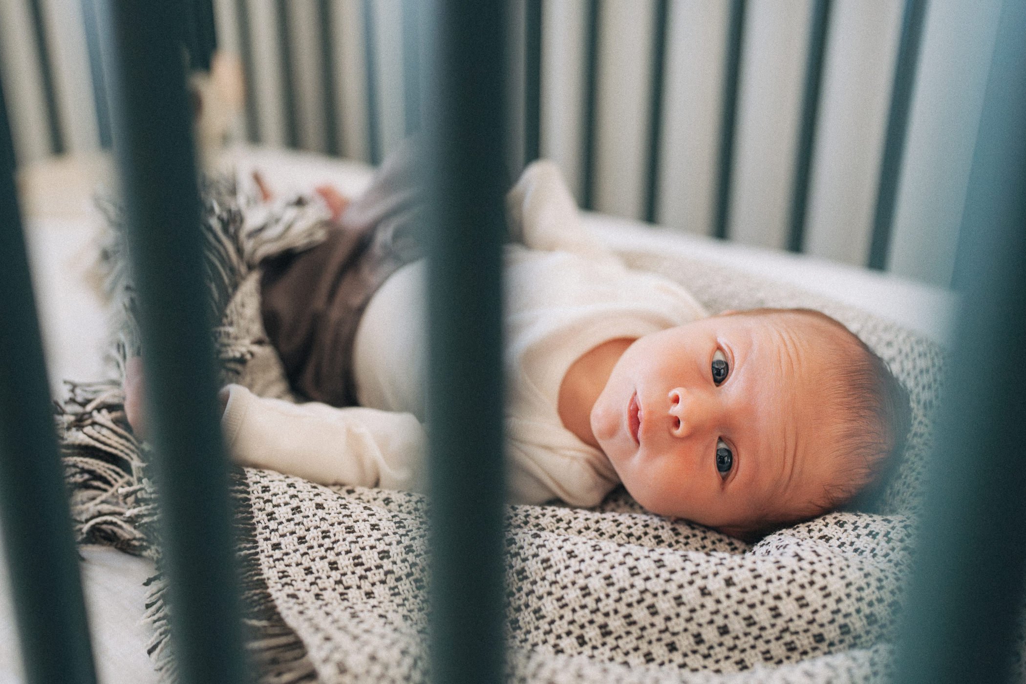 A baby lying on its side in a crib, looking out through the bars. The baby is wearing a white outfit and is resting on a patterned blanket.