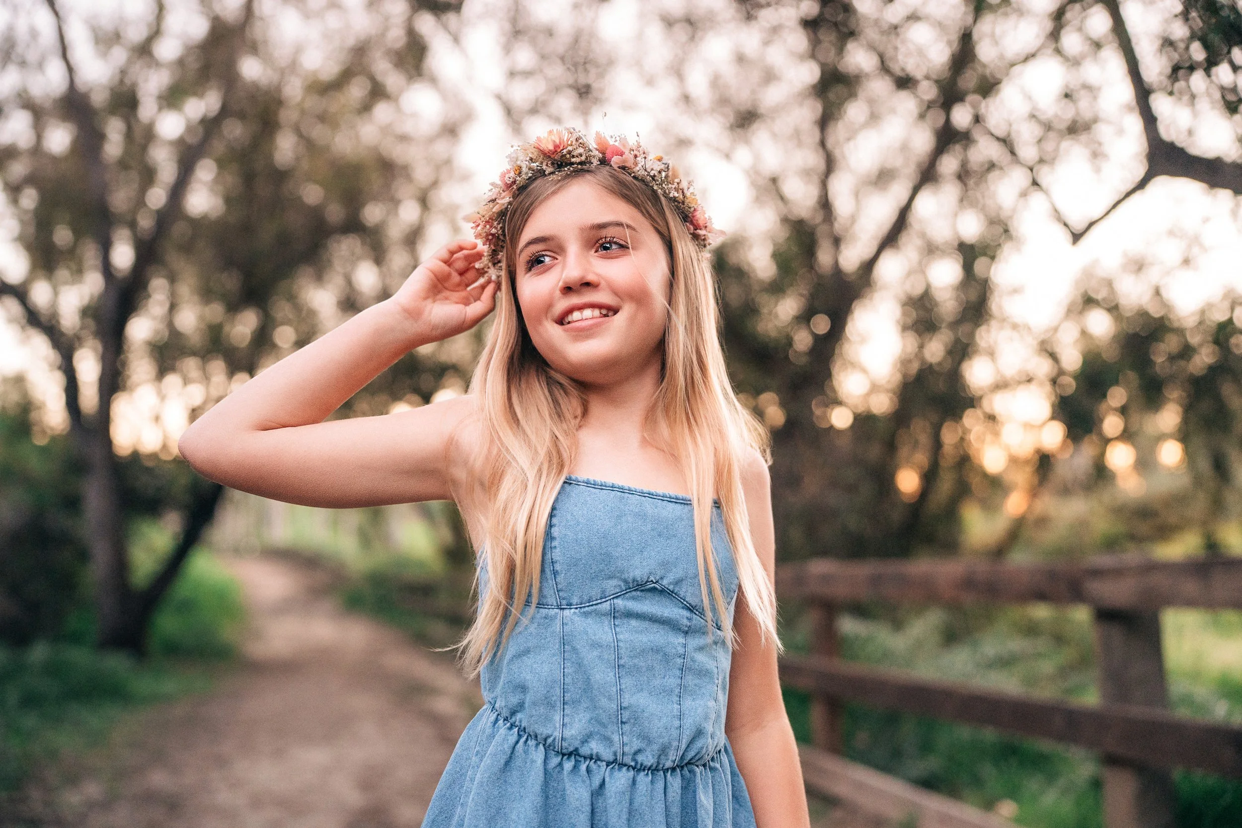 A young girl with long blonde hair wearing a denim dress and a flower crown, standing on a nature trail with trees in the background during sunset.