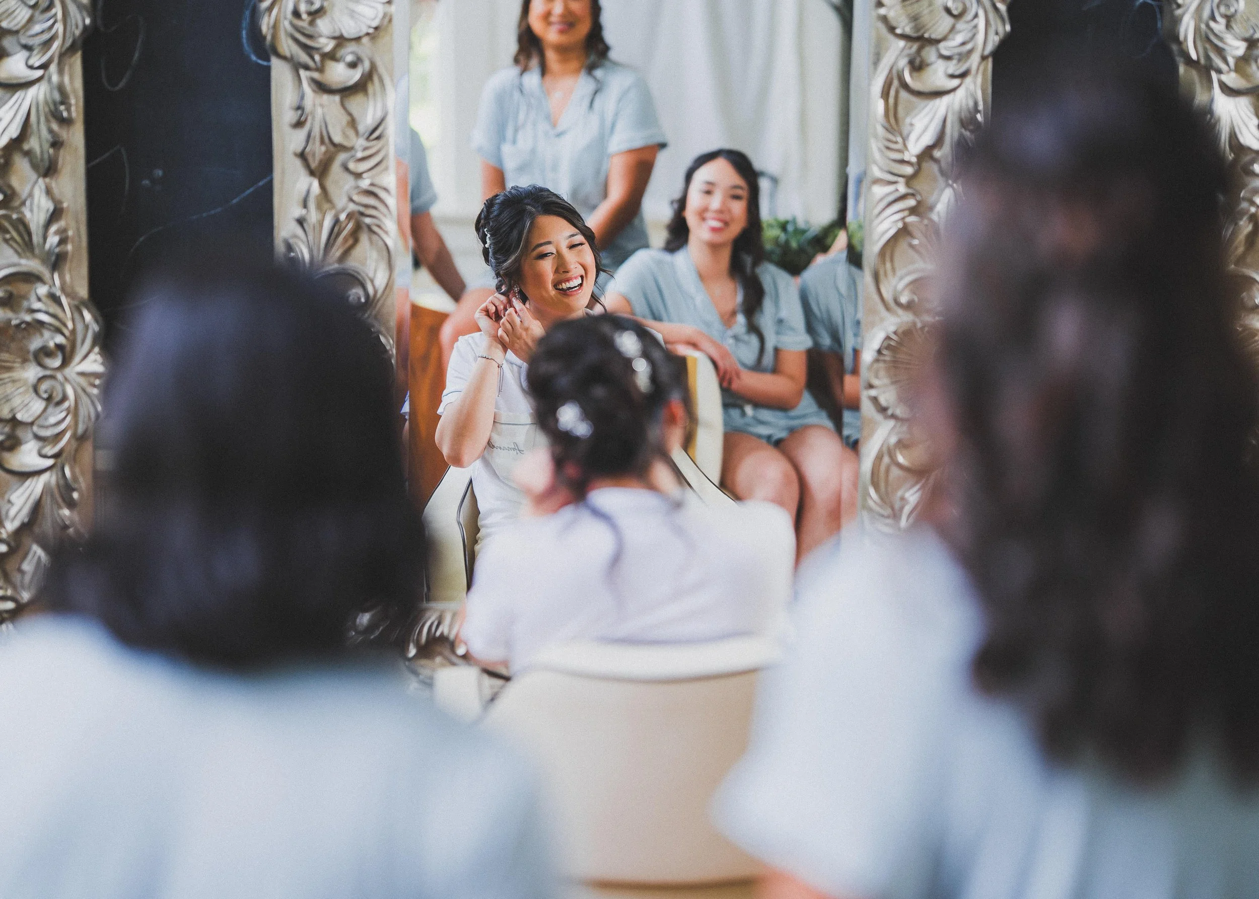 Women in matching blue outfits gathered in a room, with one woman smiling and adjusting her earring in front of a large decorative mirror, while others look on and smile.