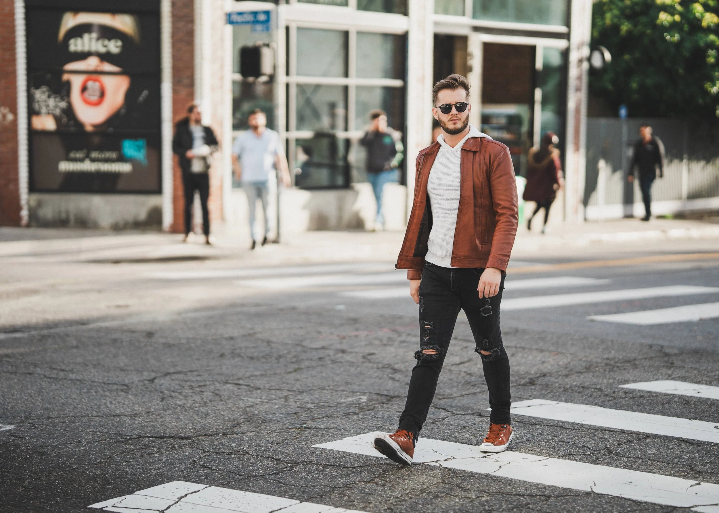 A young man with sunglasses, wearing a brown leather jacket, white sweater, ripped black jeans, and brown shoes, crossing a street with a pedestrian crosswalk, in an urban setting with people in the background.
