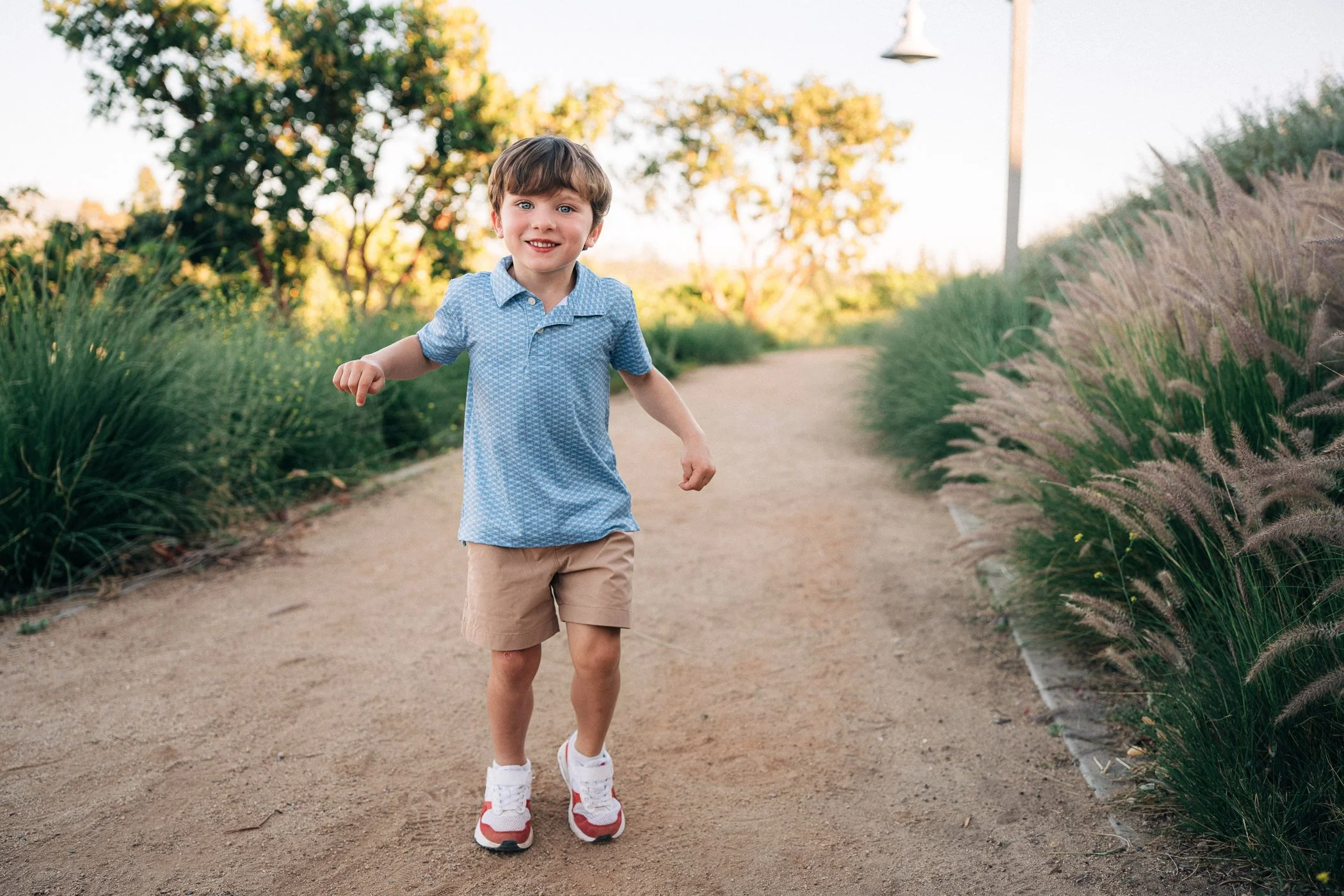 A young boy running on a dirt path in a park with trees and plants around him, smiling and wearing a blue polo shirt, beige shorts, and white sneakers.