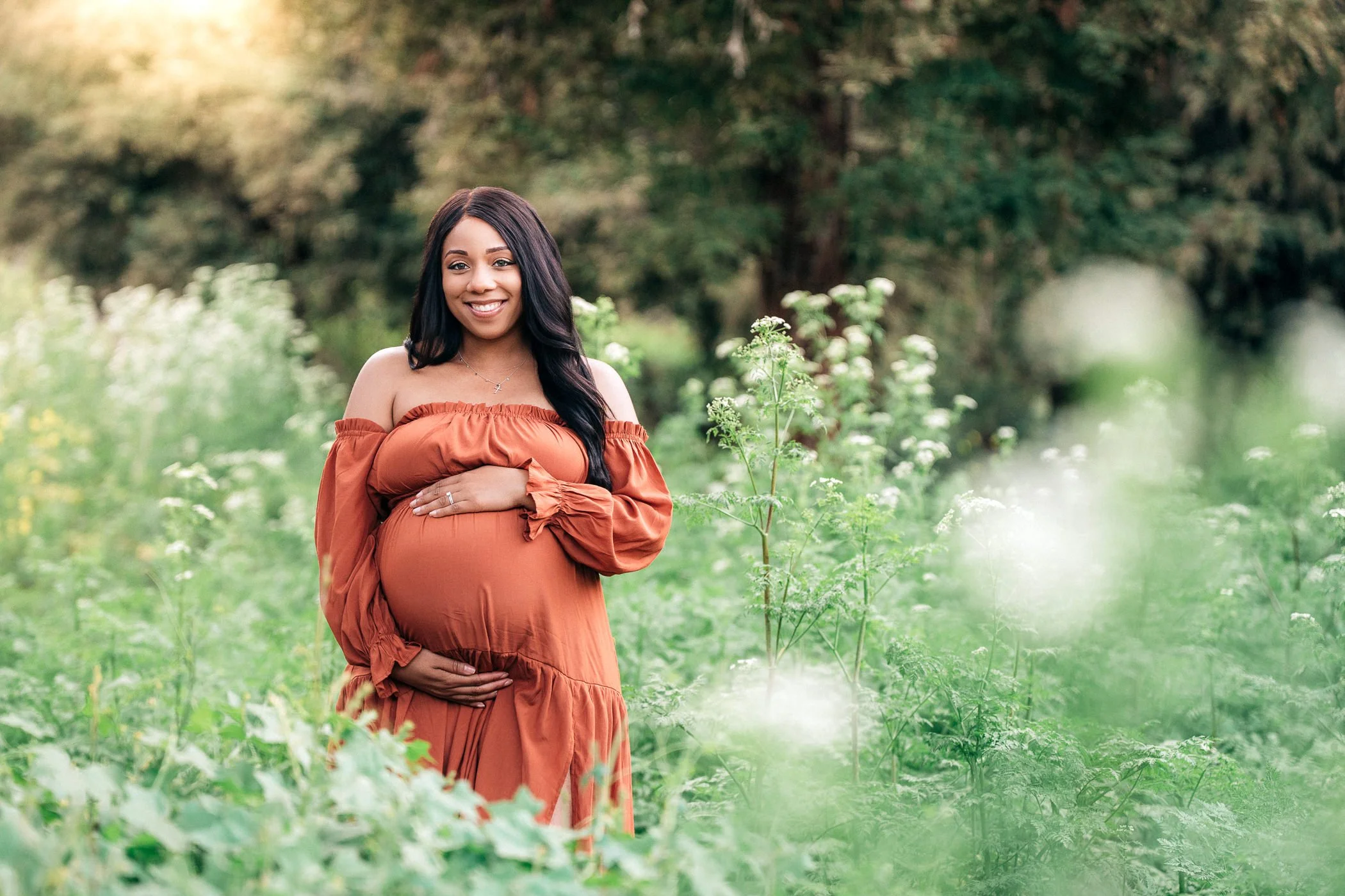 A pregnant woman in an off-the-shoulder rust-colored dress standing in a lush green garden, smiling while gently holding her baby bump.