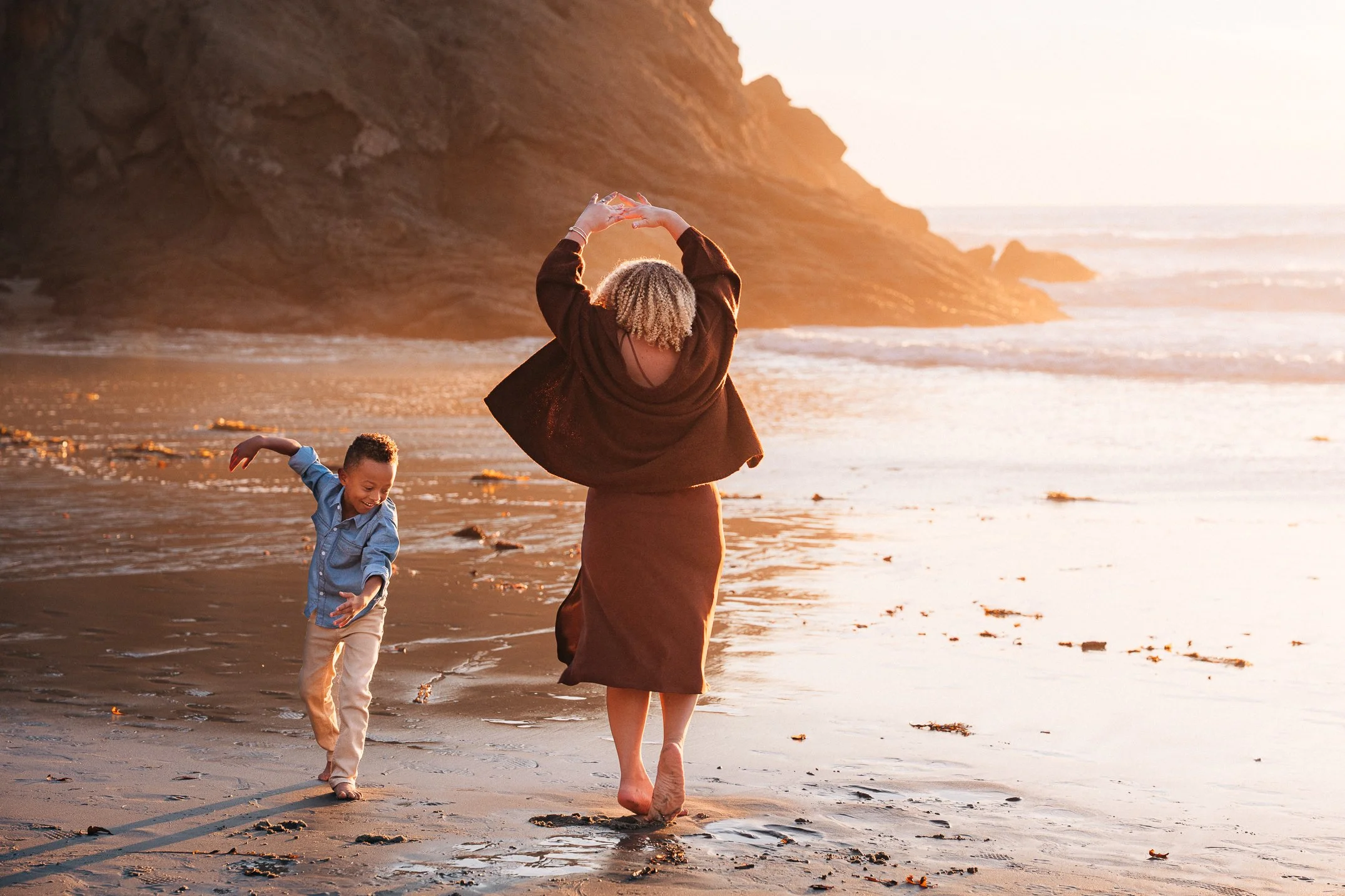 Mom dancing with arms overhead at the shoreline, son running to keep up