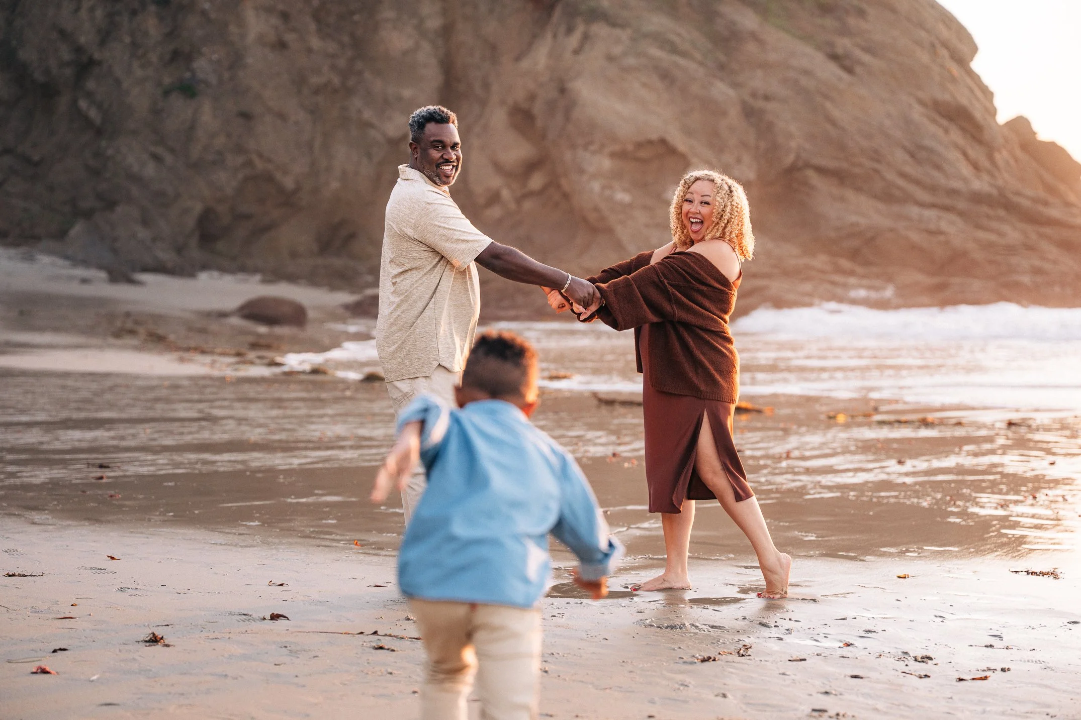 The couple holding hands by the surf as their son runs to join them