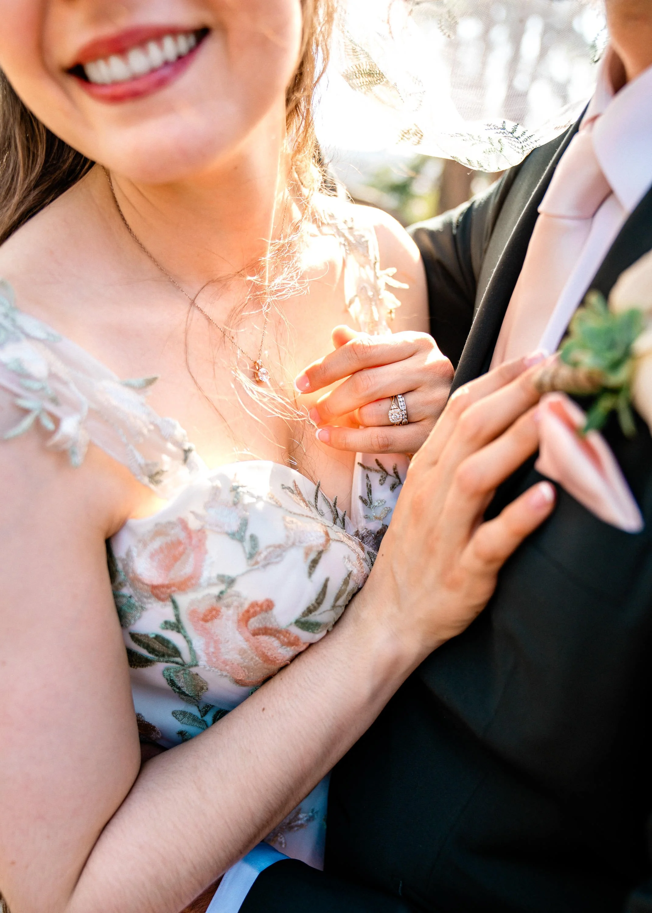 Close-up of a smiling bride and groom on their wedding day, showing the bride's floral dress, jewelry, and wedding rings, with the groom in a black tuxedo, outdoors with sunlight filtering through trees.