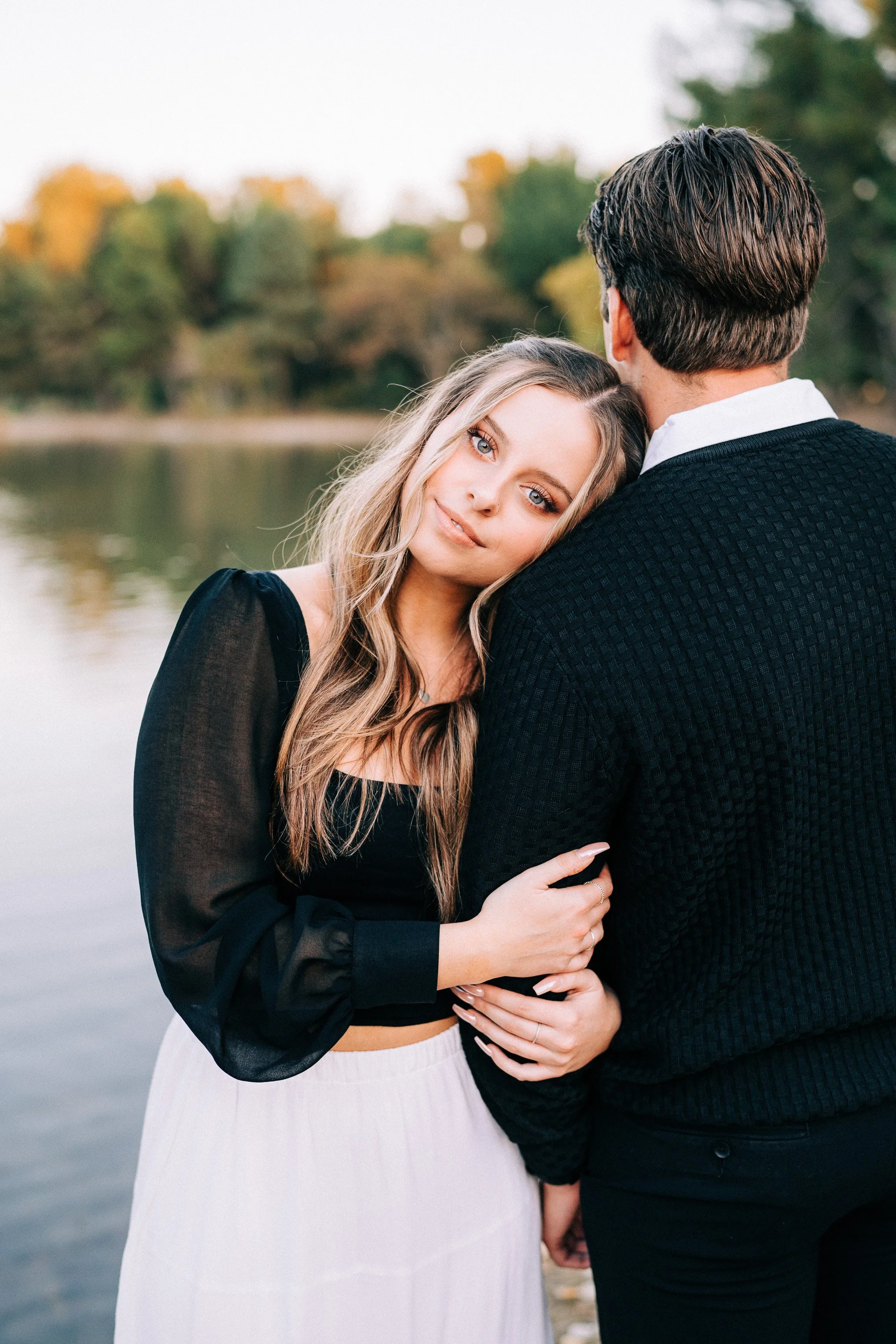 A young woman with long blonde hair and blue eyes leans her head on a young man's shoulder by a lake during autumn.
