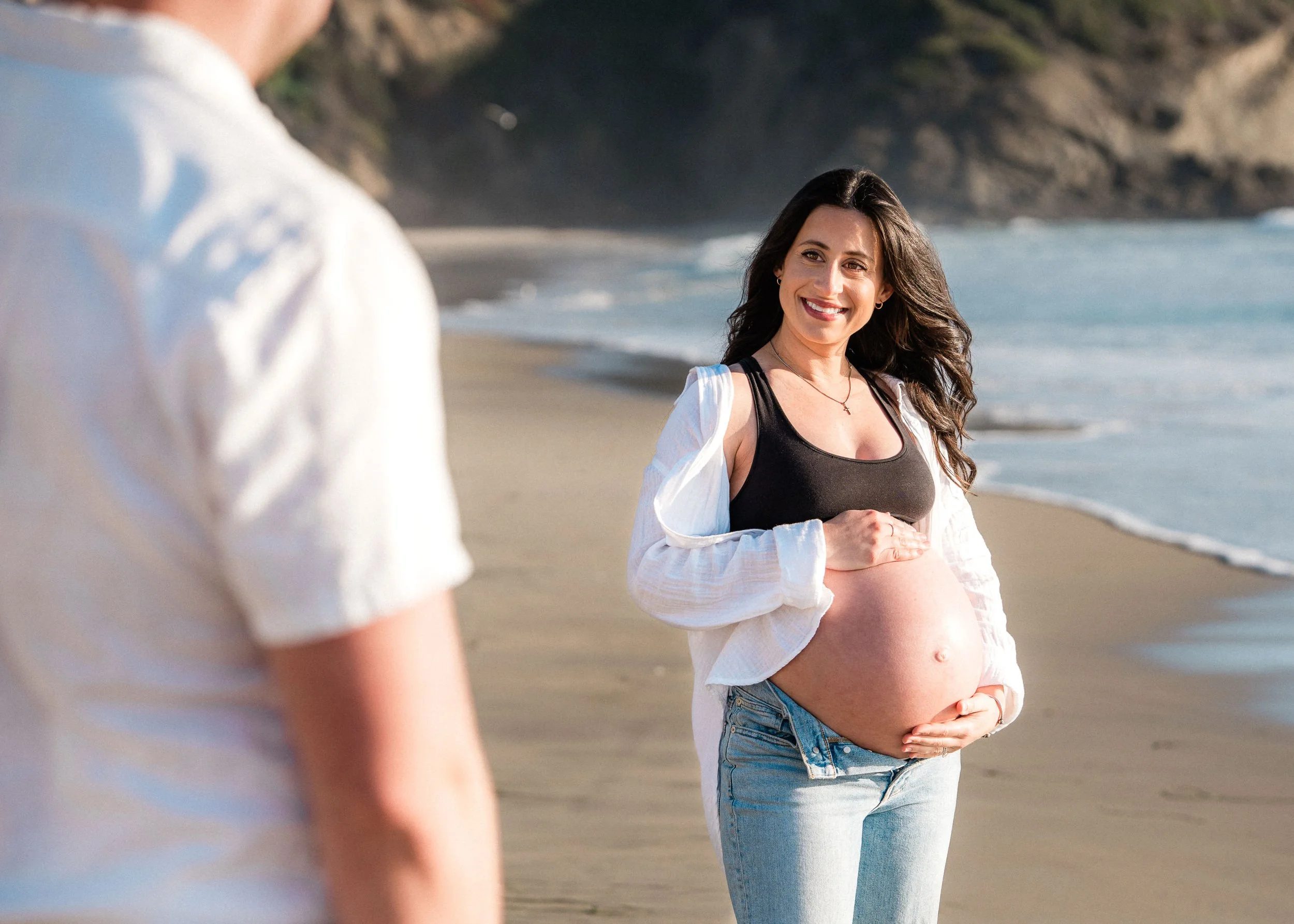 A pregnant woman with long dark hair smiling on a beach, holding her belly, while a man in a white shirt stands nearby.