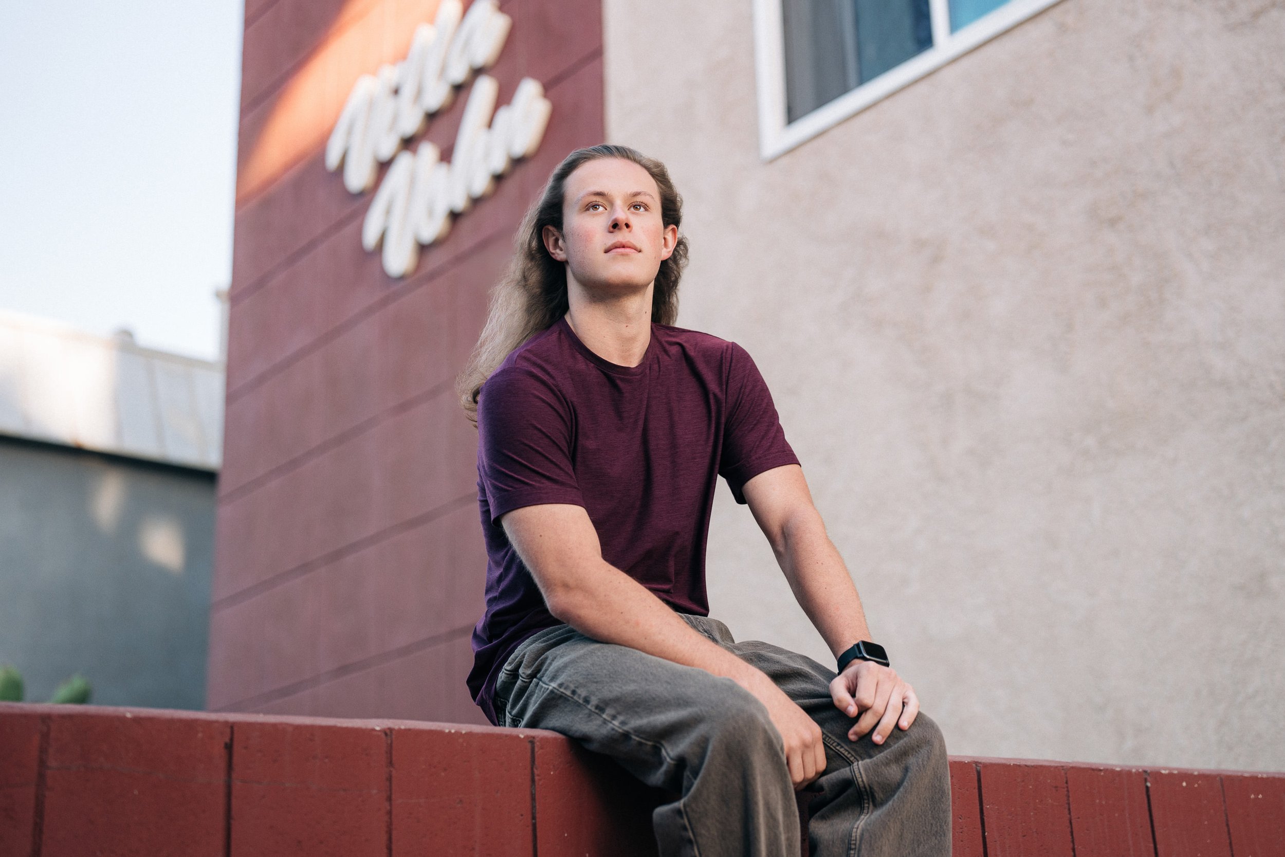 Young man with long hair sitting on a red brick ledge outside a building with a beige wall and large window, wearing a maroon t-shirt, gray pants, and a smartwatch.