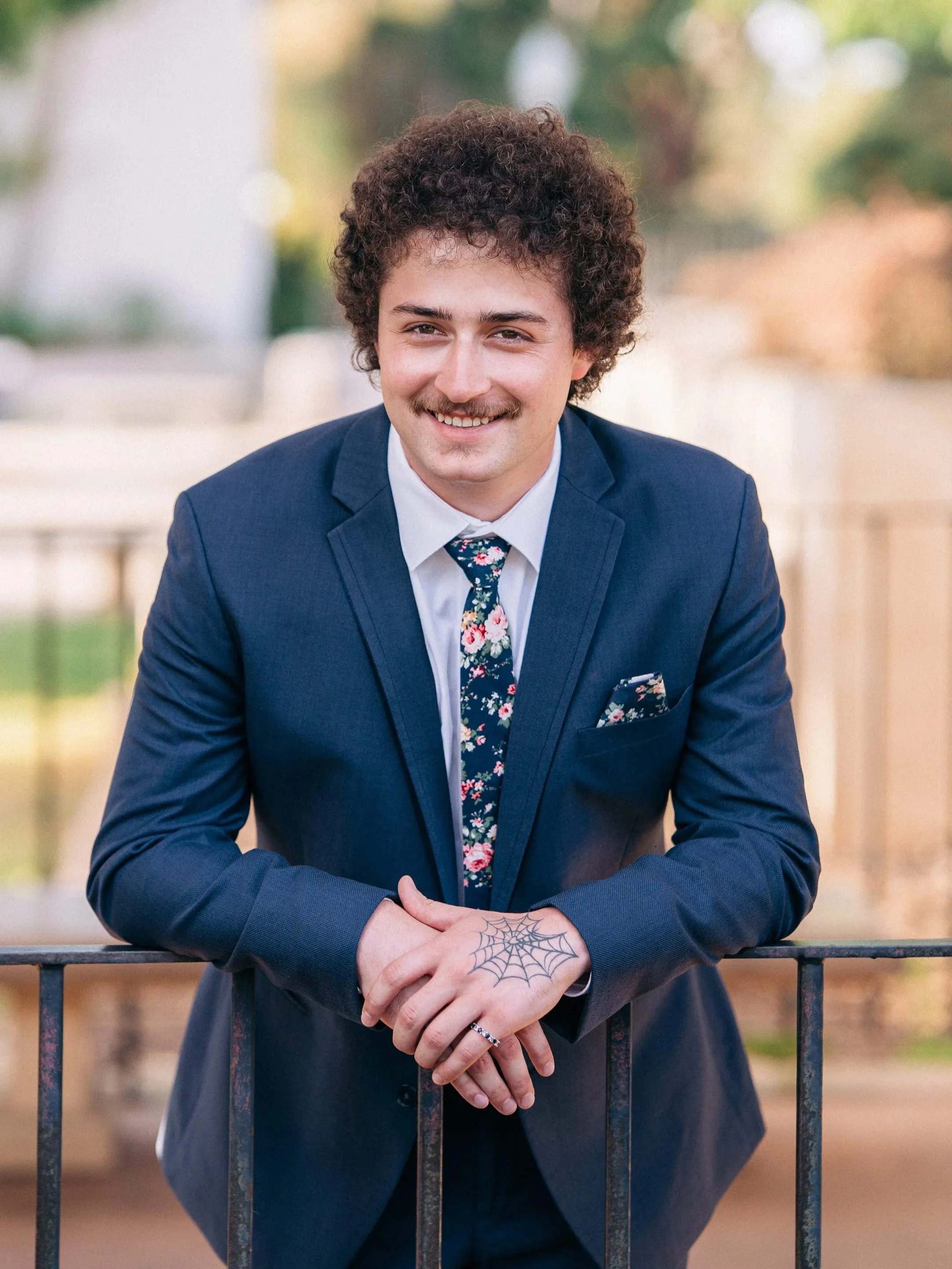 A young man with curly hair, a mustache, and a cheerful expression, wearing a blue suit, floral tie, and pocket square, leaning on a railing outdoors.