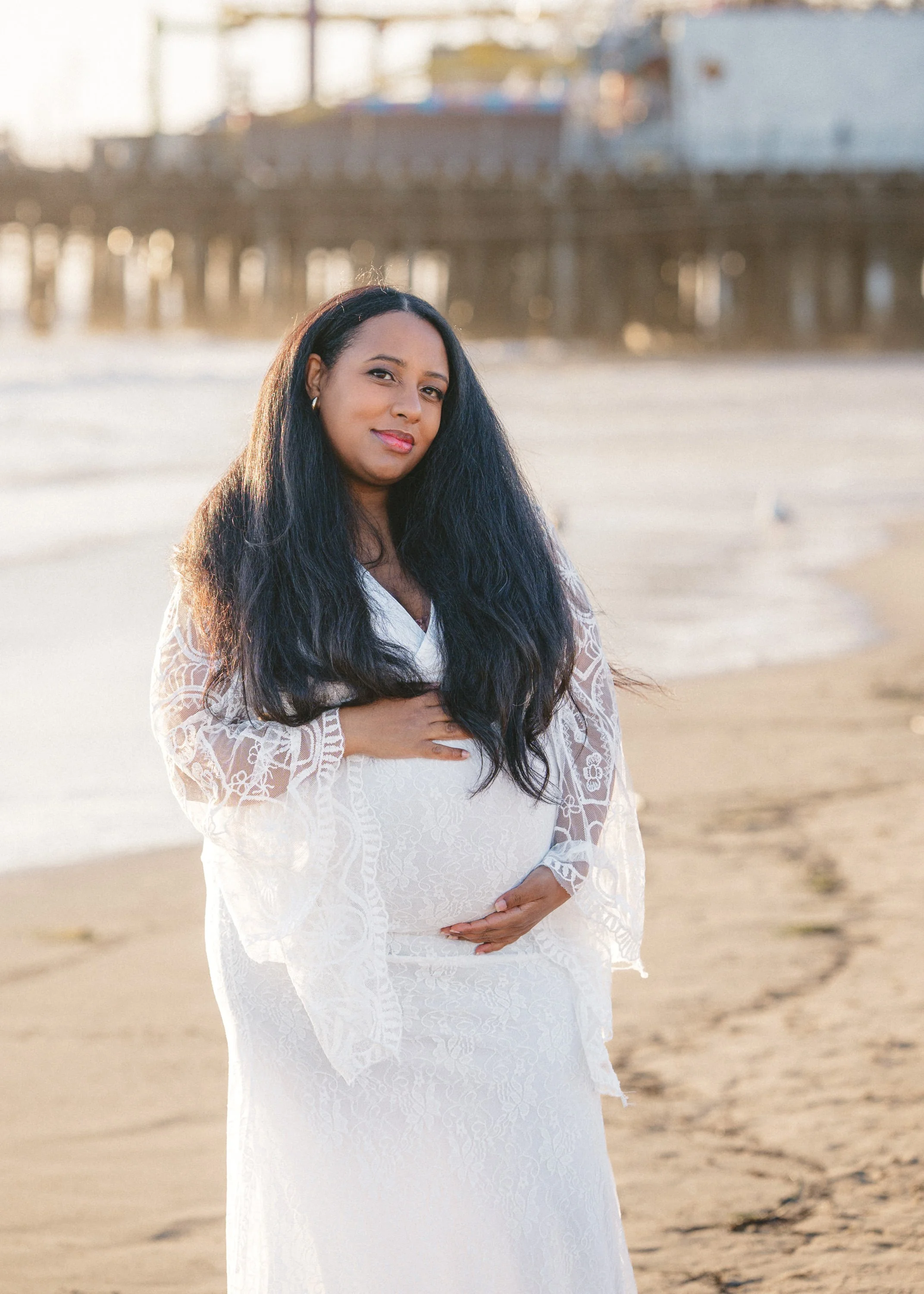 A pregnant woman in a white lace dress standing on a beach with her hand on her belly, facing the camera. In the background, there are beach structures and a pier, with sunlight illuminating the scene.