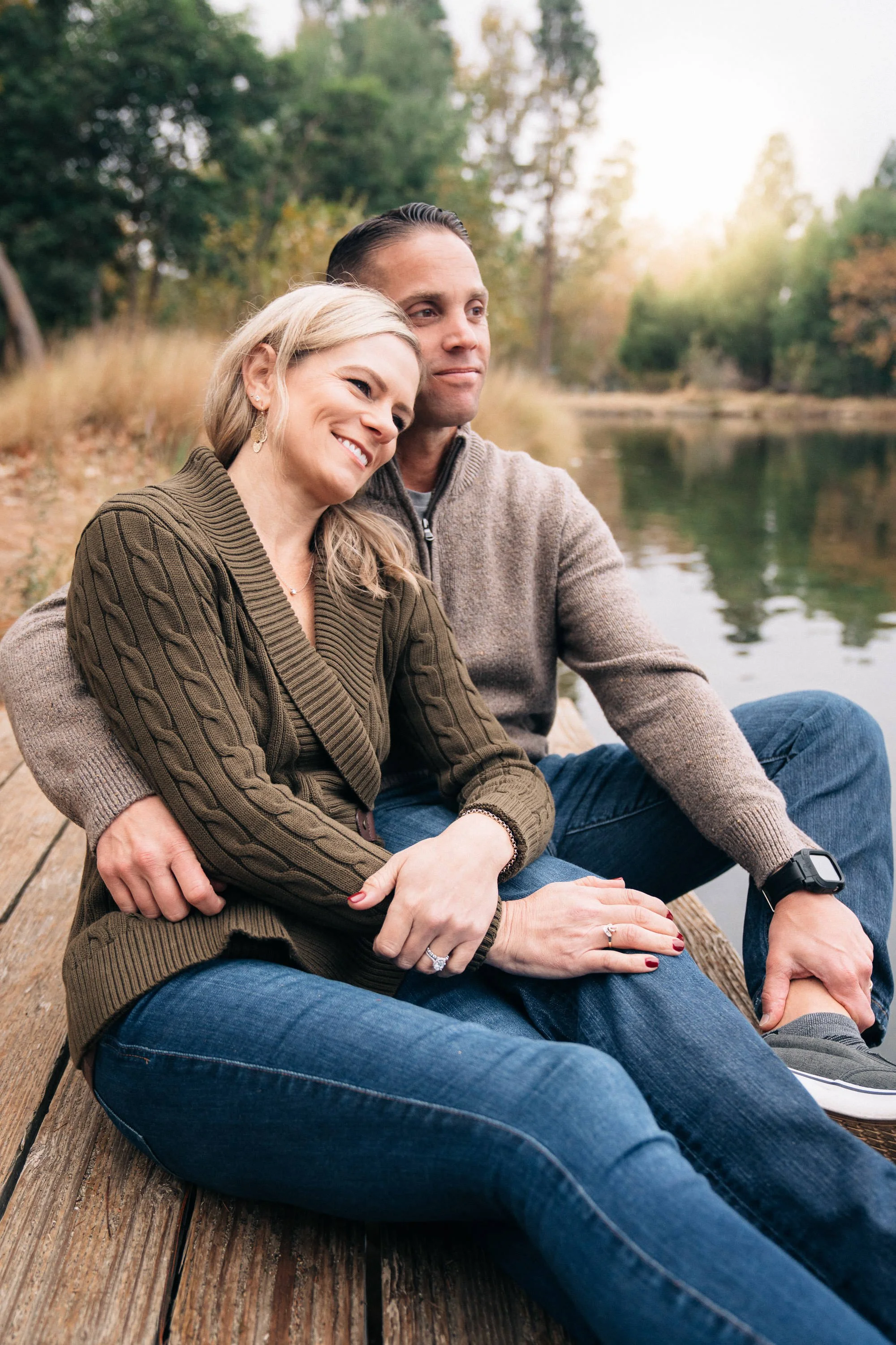 A happy couple sitting on a wooden dock by a lake during autumn, embracing and smiling.