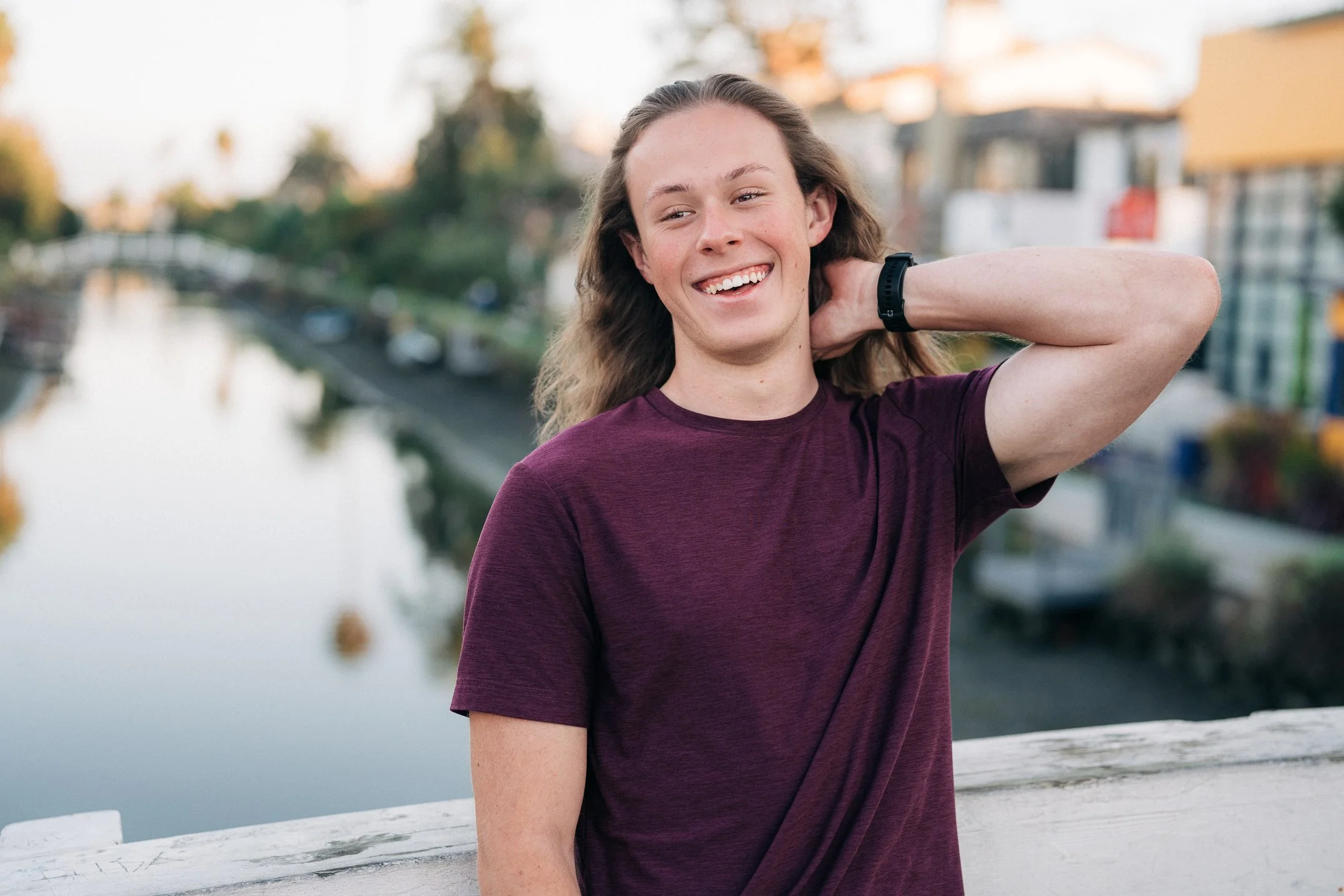 A young man with long wavy hair, wearing a maroon T-shirt and a black watch, smiling and touching the back of his neck while sitting outdoors near a body of water with buildings and trees in the background.