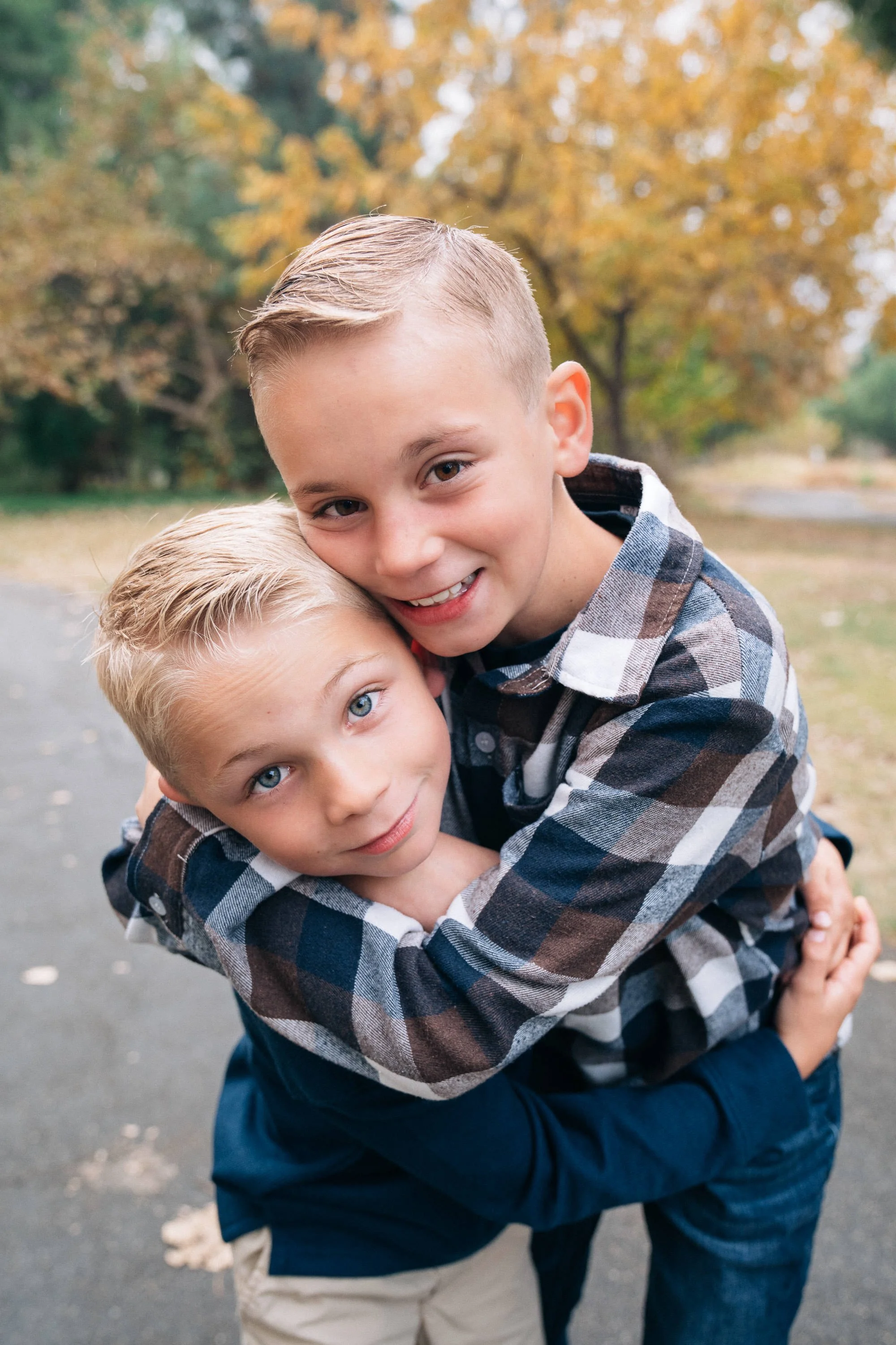 Two young boys hugging each other outdoors during fall, with trees showing orange and yellow leaves in the background.