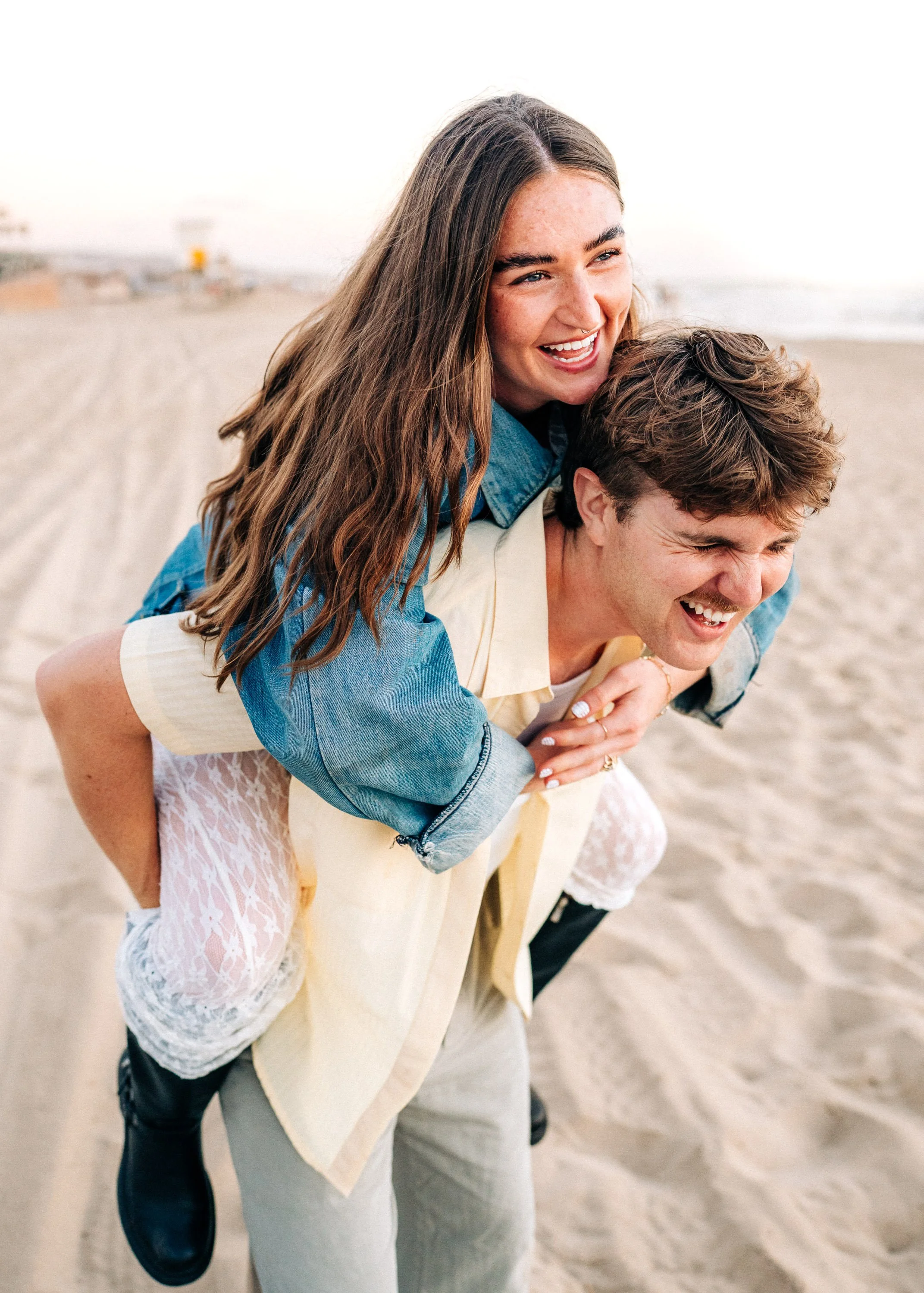 A young woman giving a piggyback ride to a young man on the beach, both smiling and enjoying themselves.